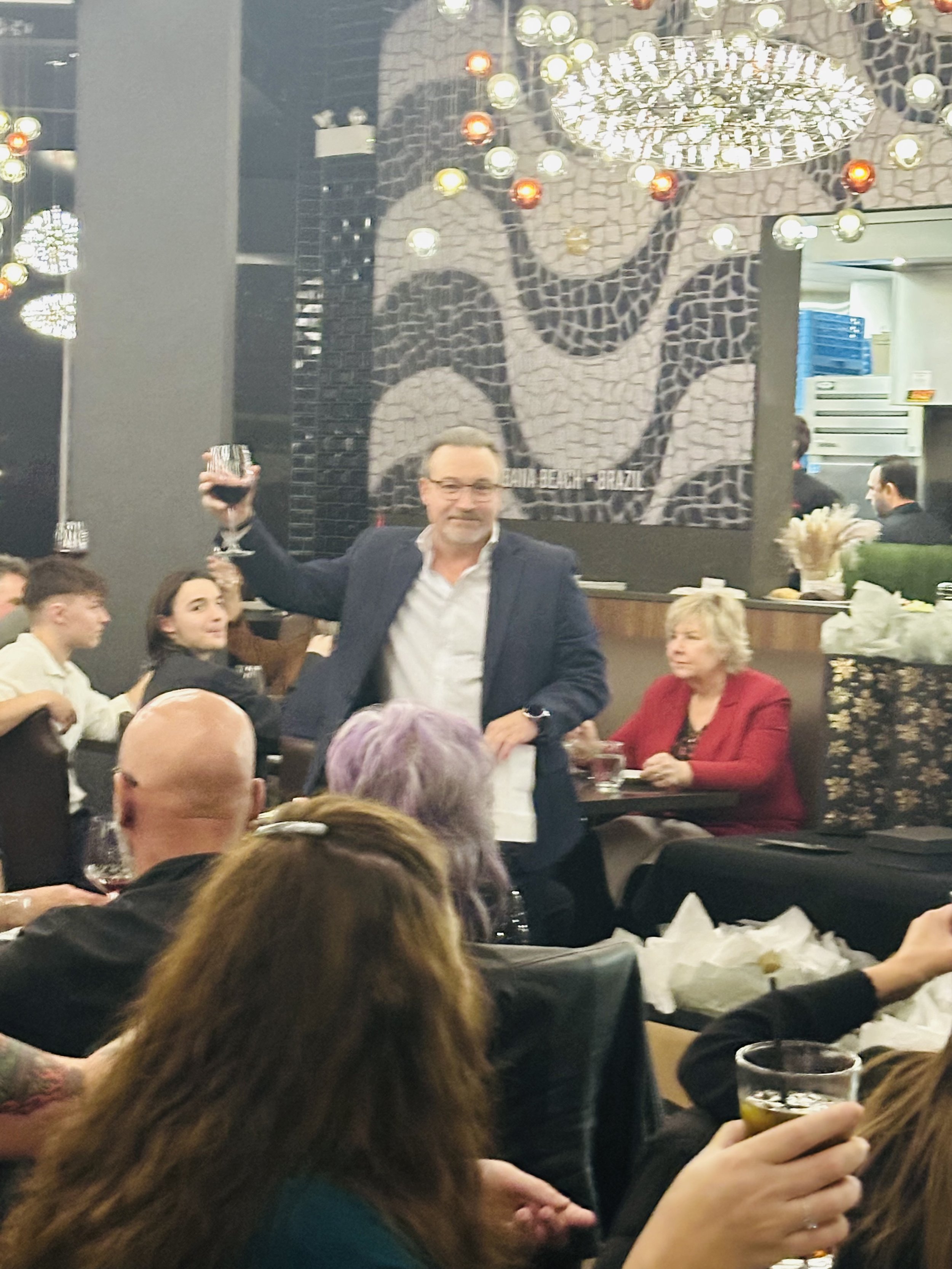 A man in a dark suit holding a glass of red wine while standing at a table in a crowded restaurant or bar. People seated around him are engaged in conversation, some holding drinks. The background features a large mosaic wall art, modern lighting fix