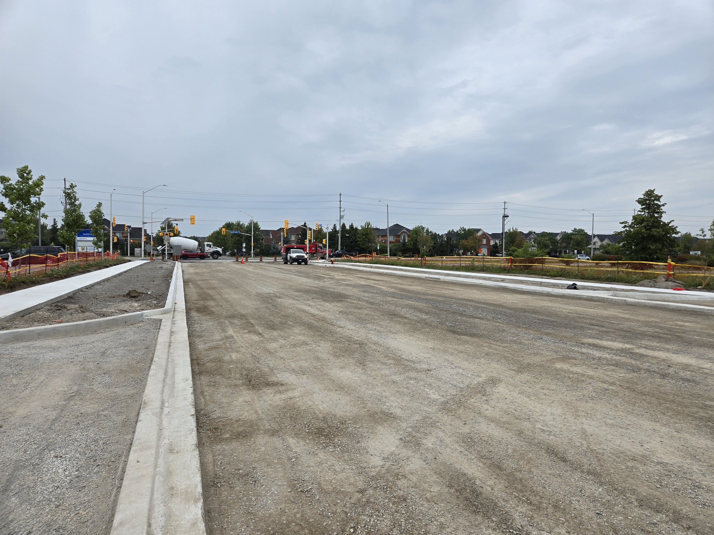 Road construction site with partially paved road, construction cones, and vehicles, with houses and traffic lights in the background on a cloudy day.