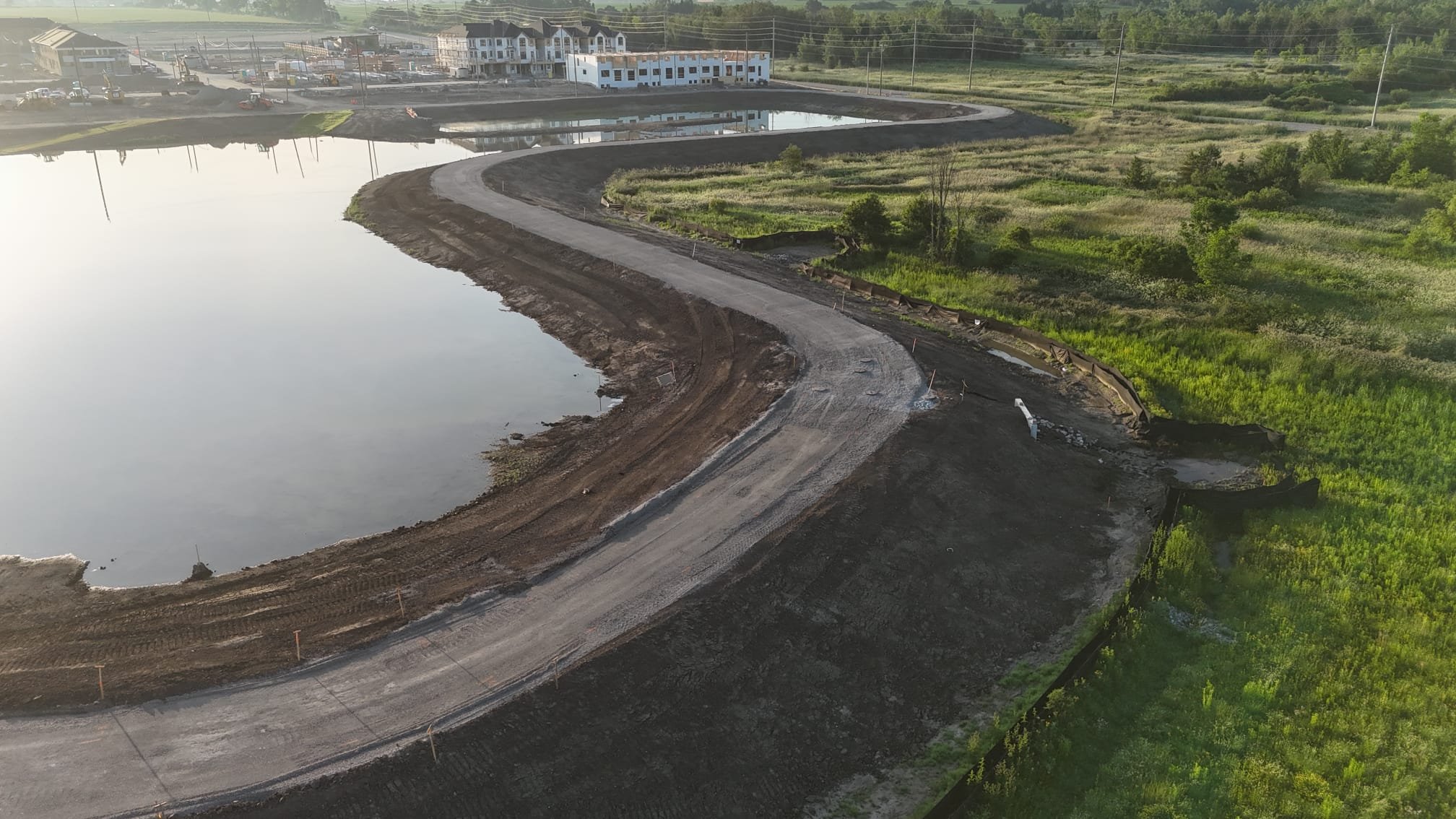 An aerial view of a construction site with a partially built road curving around a body of water, with equipment and materials along the edge, and green vegetation nearby.