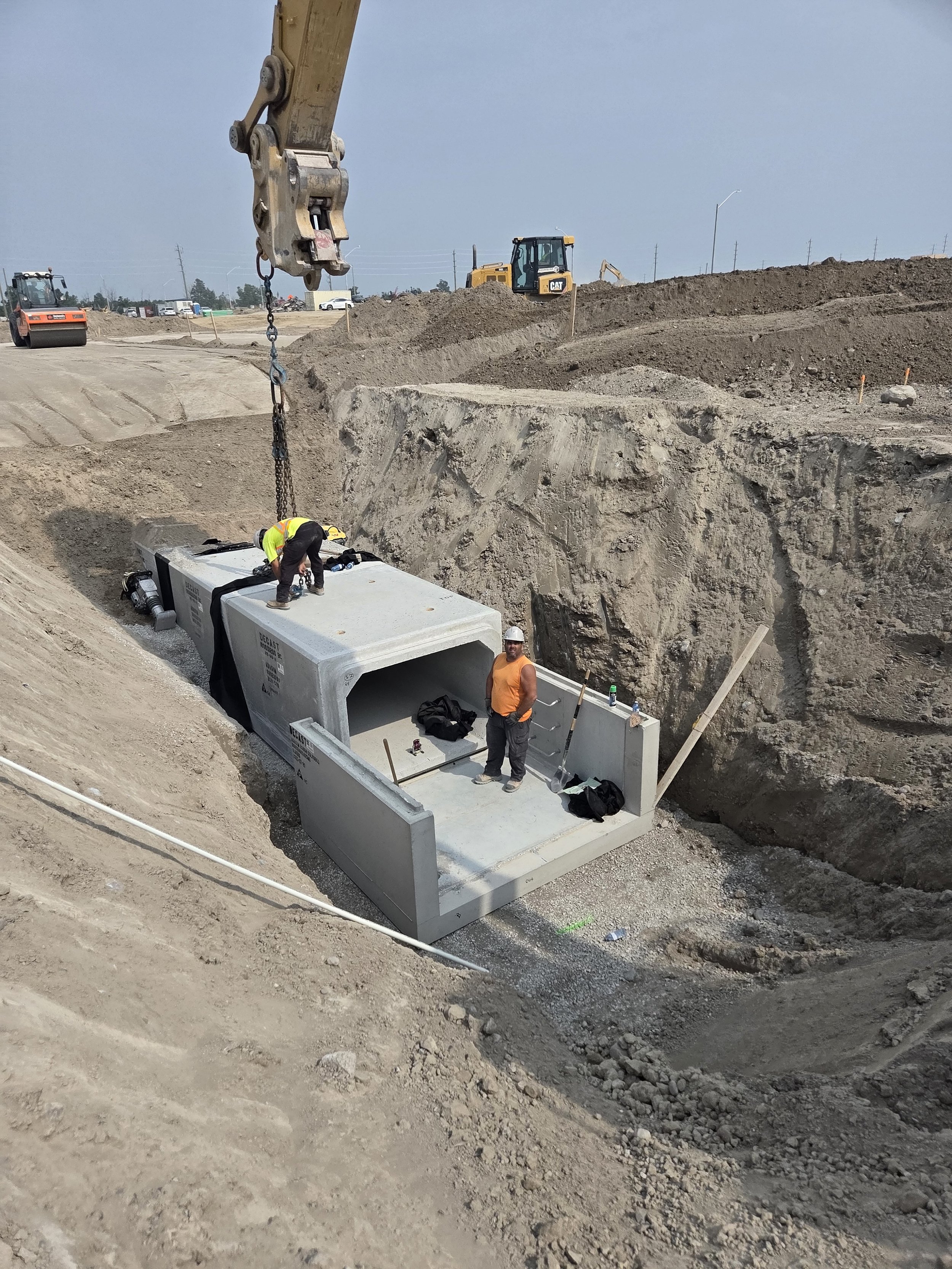 Construction workers installing a large concrete culvert pipe in a trench at a construction site, with construction equipment and dirt mounds in the background.