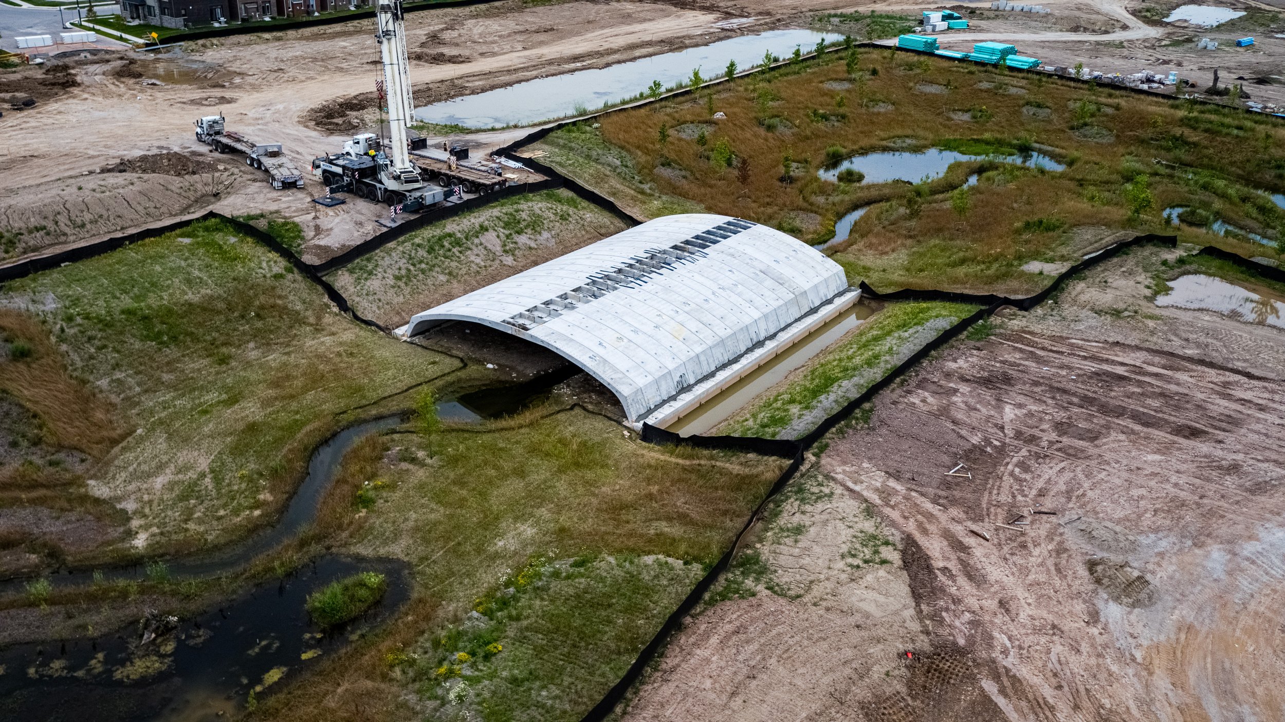 A construction site with a large white greenhouse structure, construction equipment, and landscaped areas with small ponds and streams.