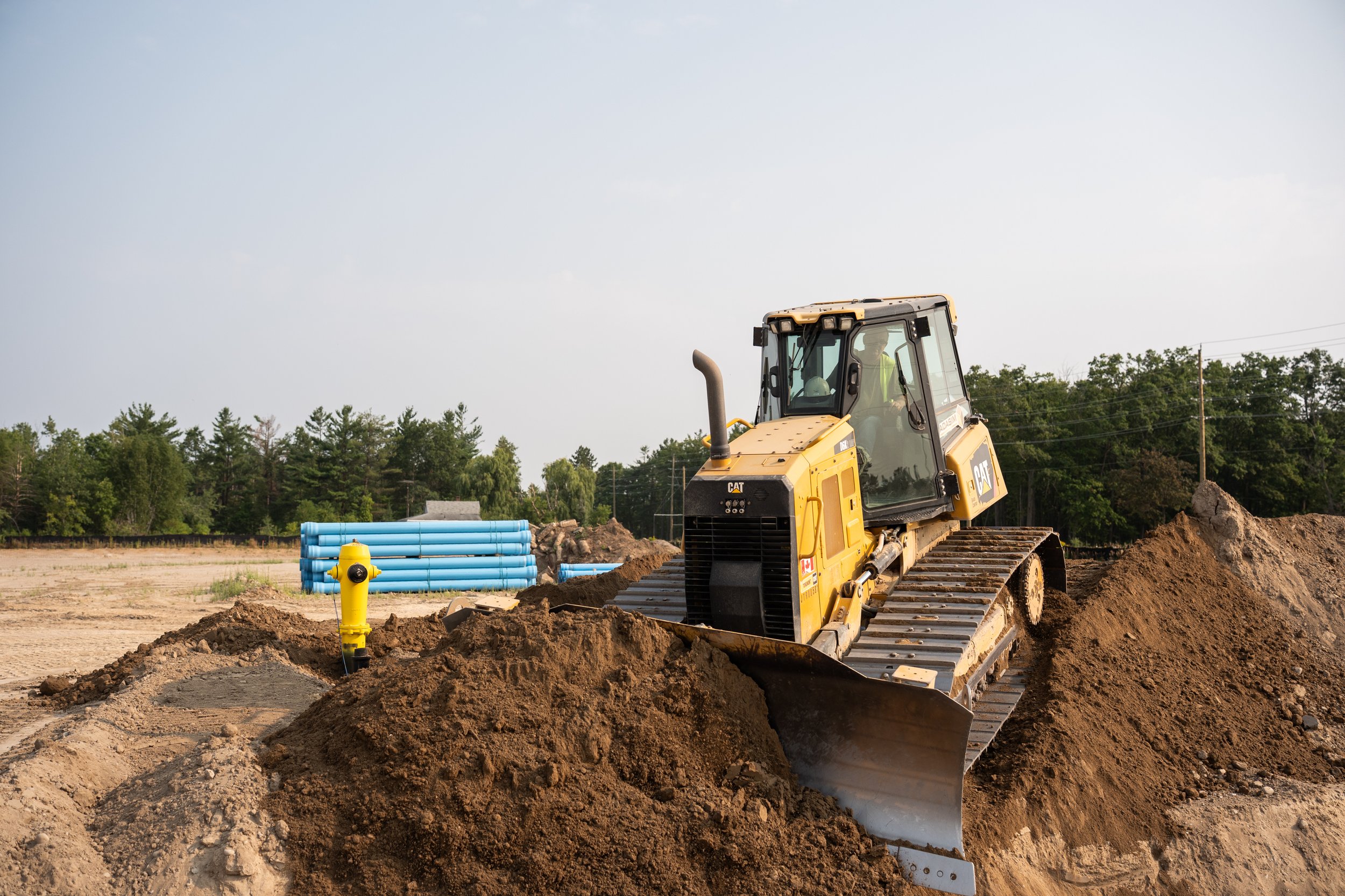 A yellow bulldozer pushing a pile of dirt on a construction site with blue pipes and trees in the background.