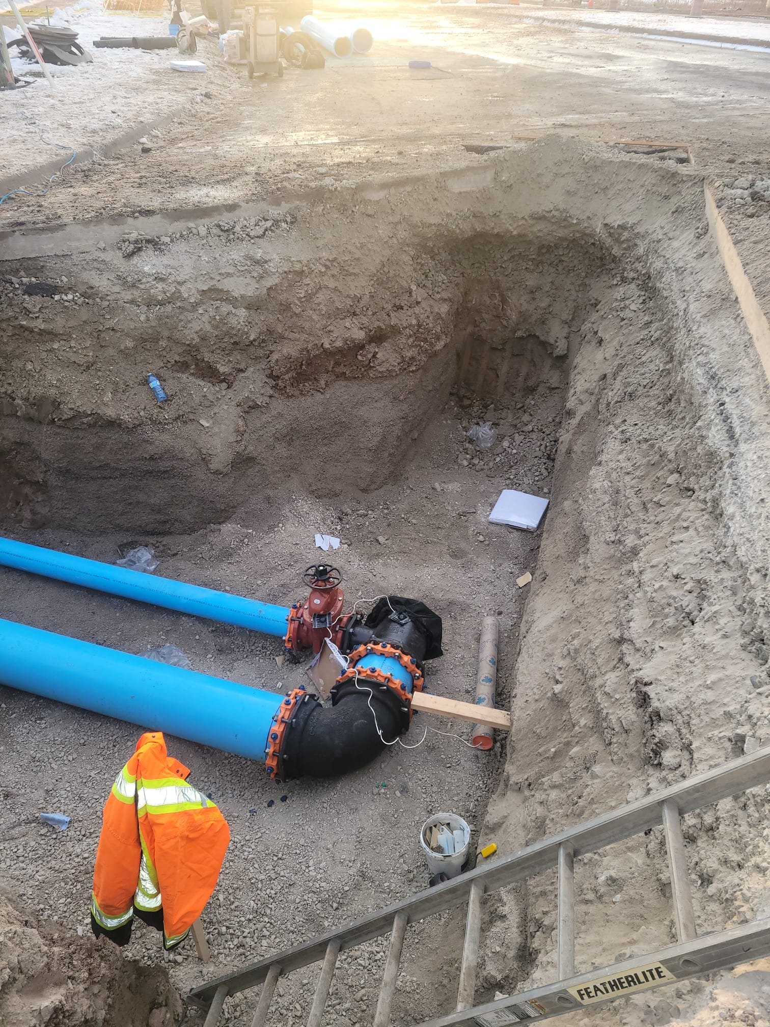 Construction site with large blue pipes and a black elbow joint, a high-visibility orange jacket hanging on a ladder, dirt excavation with tools and debris, and a ladder labeled 'Featherlite'.