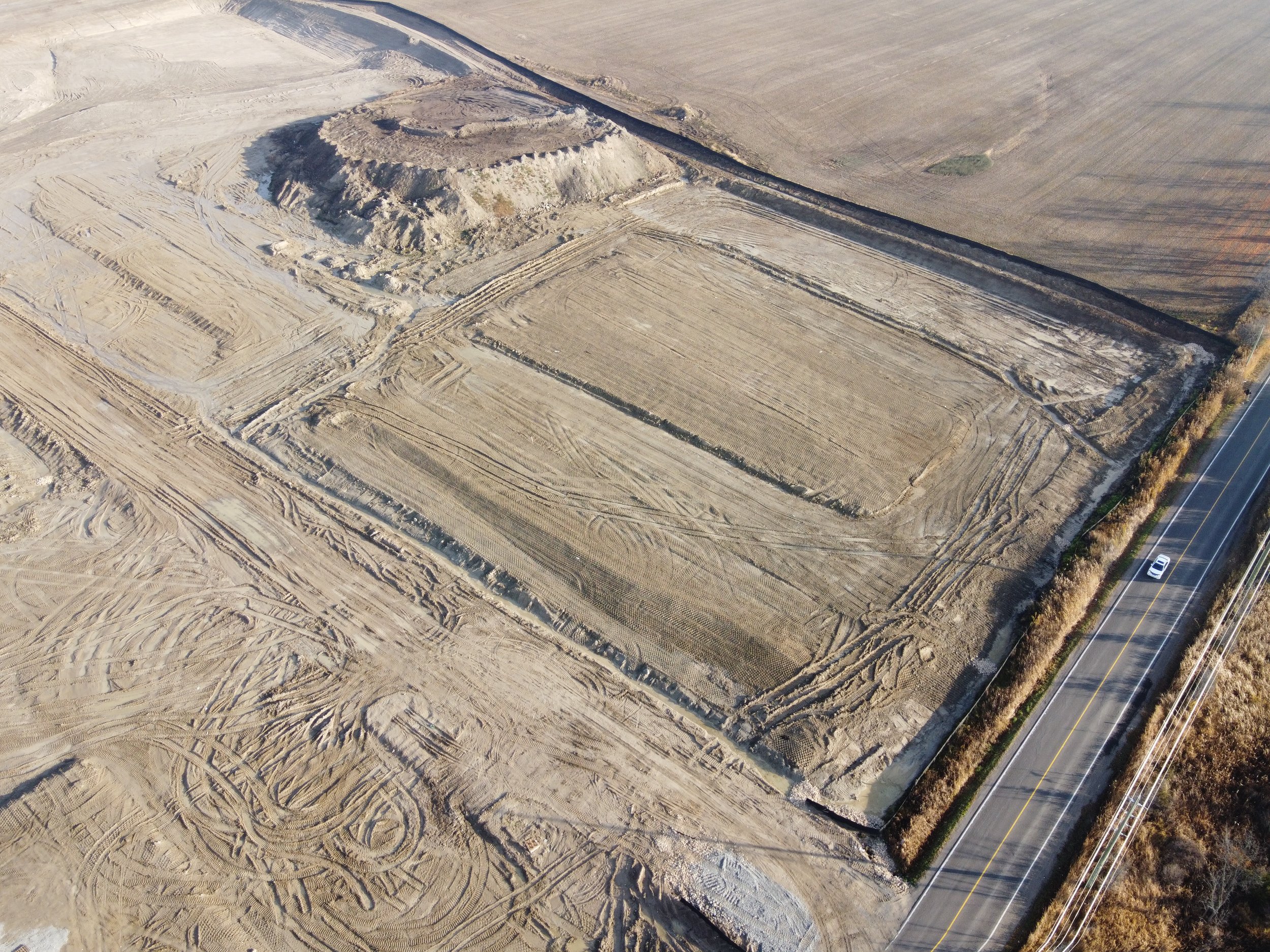 Aerial view of an empty dirt lot with excavation marks, bordered by a road on the right side and open fields on the other sides.