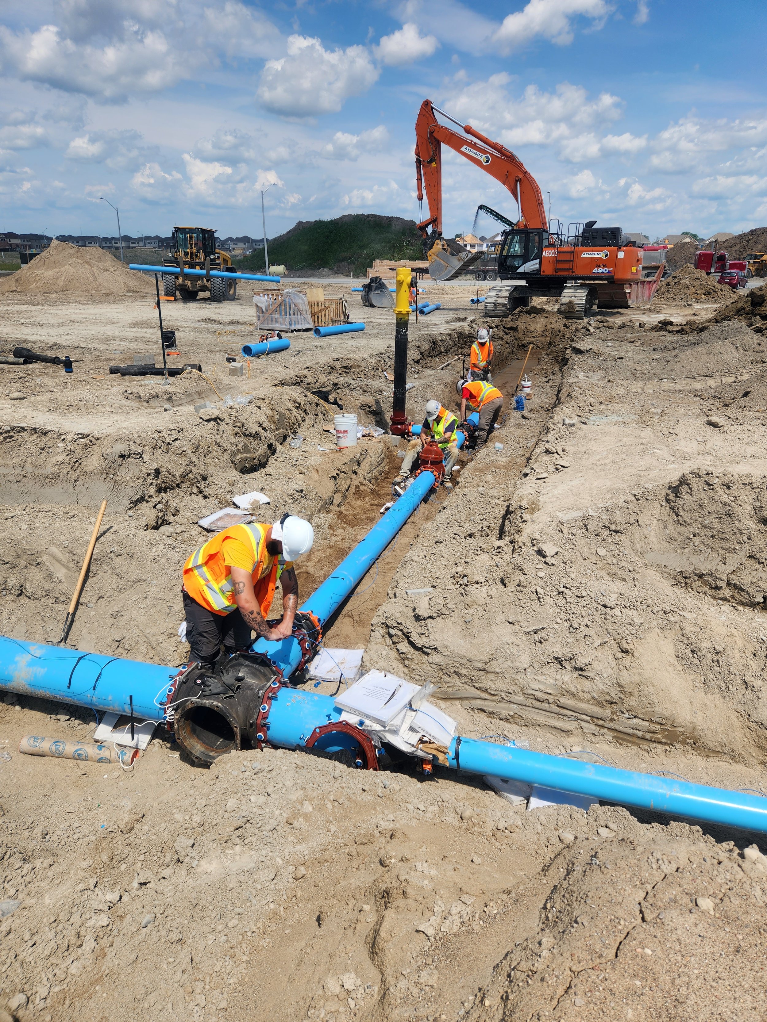 Construction workers in safety vests and helmets working on installing underground pipes at a construction site. Large construction equipment and blue pipes are visible.