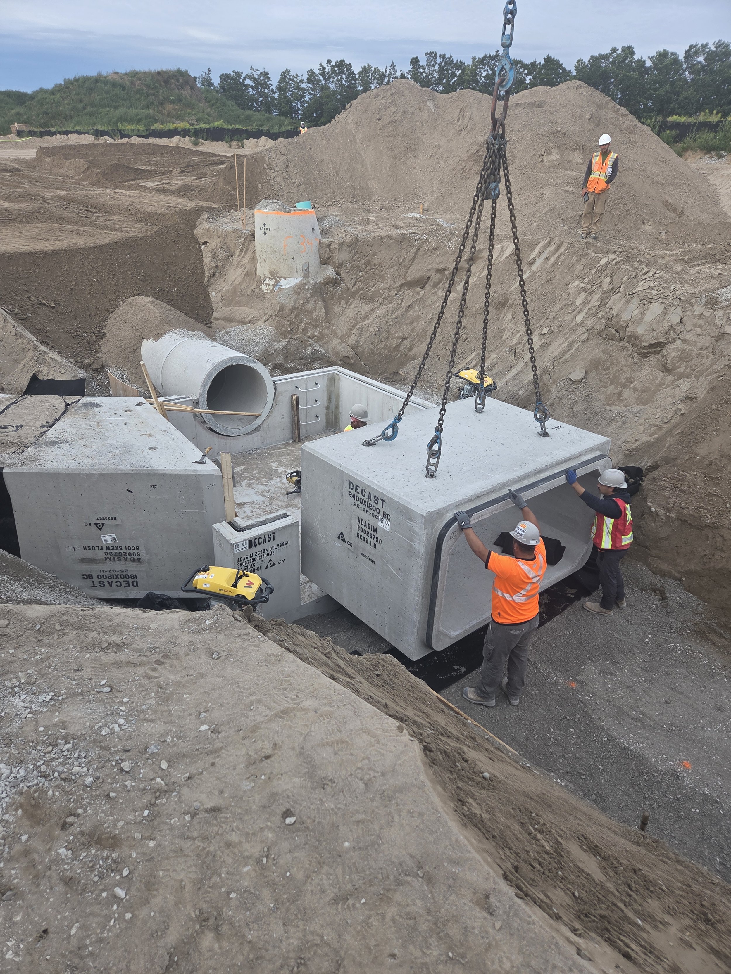 Construction workers installing large concrete utility boxes in an excavated site with dirt and gravel. One worker is guiding a concrete box while another is secured nearby, and additional workers are on the site with safety gear.