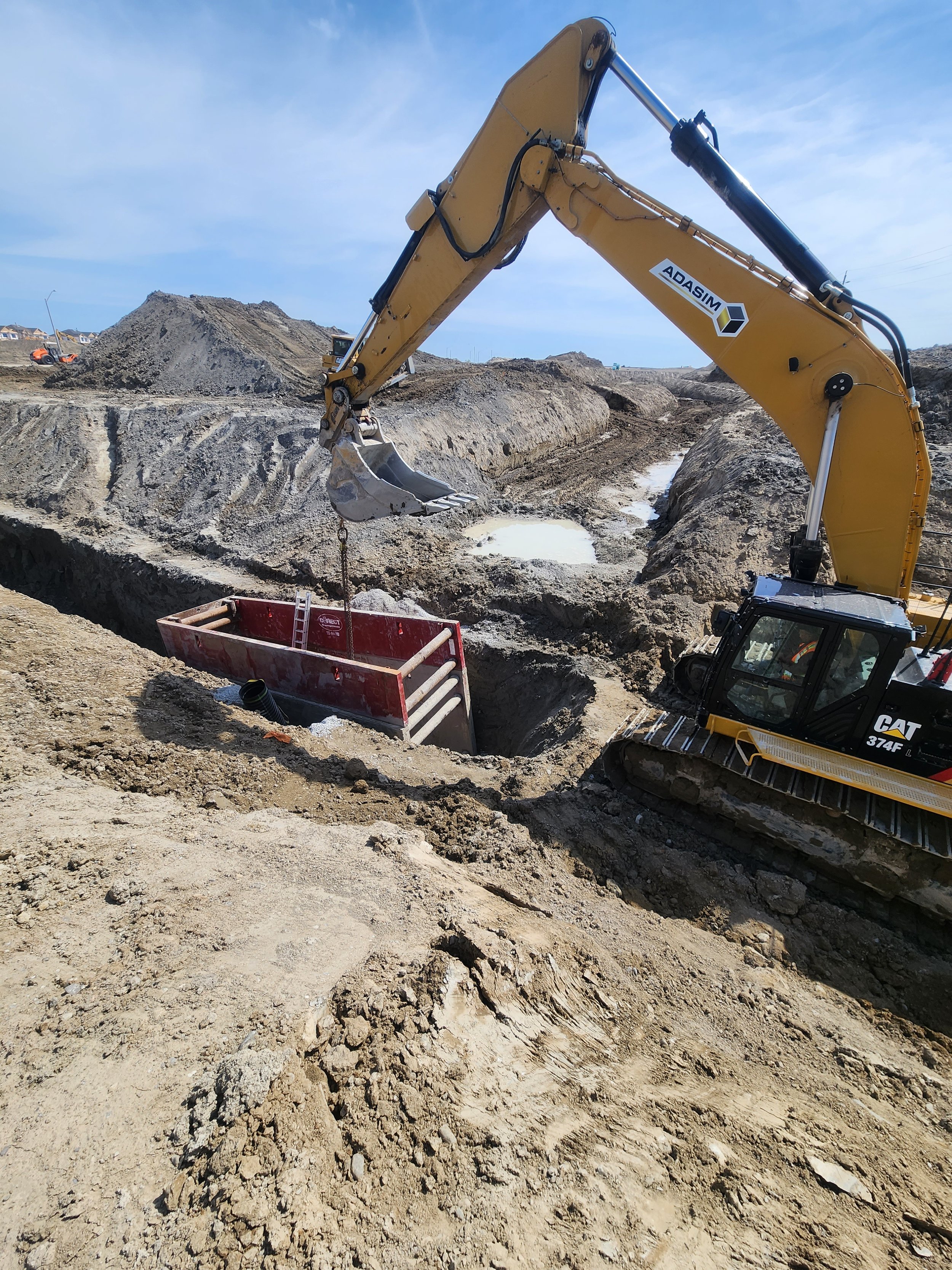 Construction site with a large yellow excavator digging in the dirt, with a red container and a ladder nearby, and dirt mounds in the background.