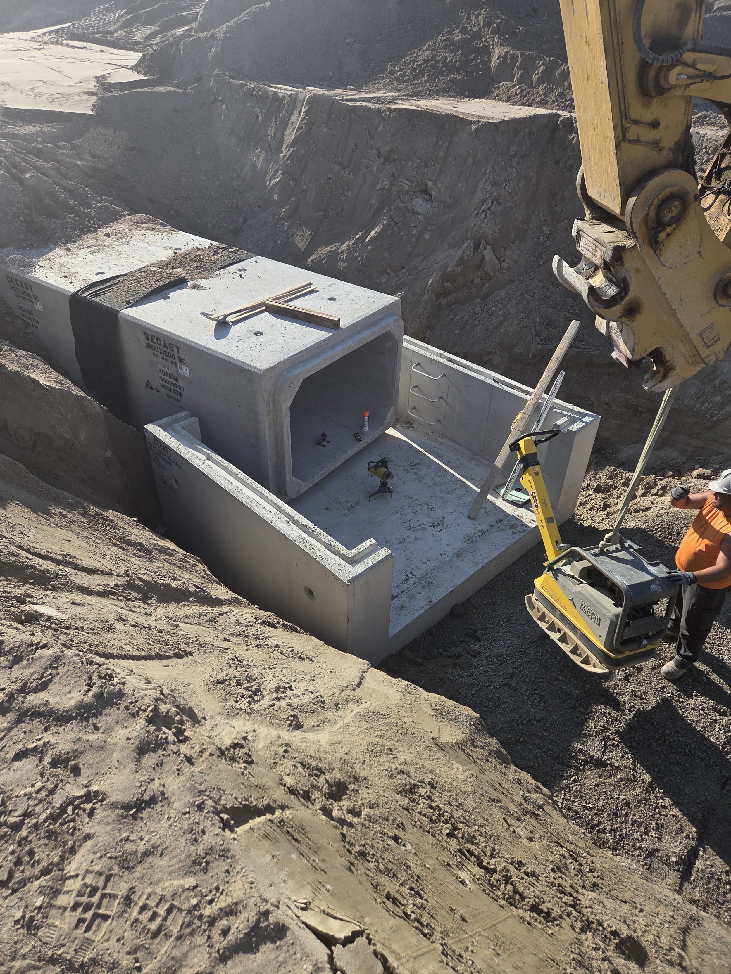 Construction worker operating heavy machinery and installing large concrete pipes in a trench at a construction site.