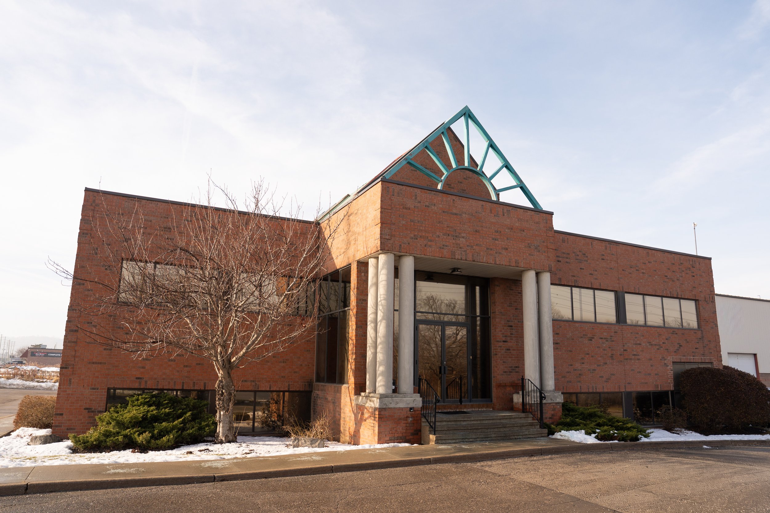 A brick commercial building with large windows, a glass door, and a decorative triangular metal structure on the roof, surrounded by a parking lot, some shrubbery, and a leafless tree in the foreground, on a sunny winter day.