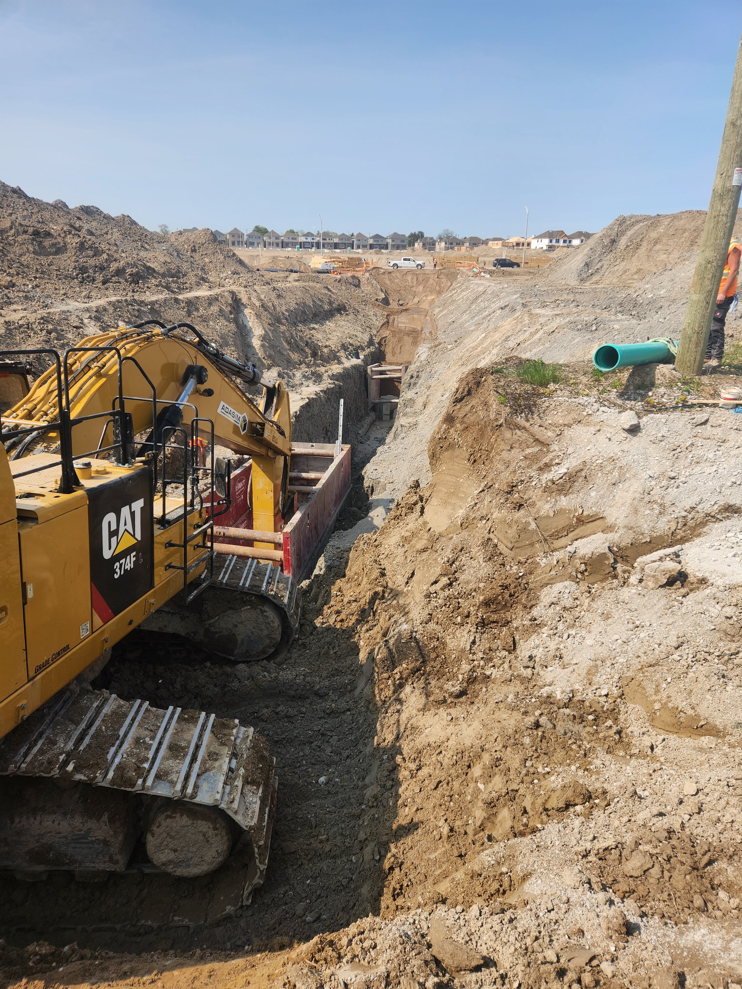 Construction site with excavator digging a trench in dirt soil, and houses in the background under clear blue sky.
