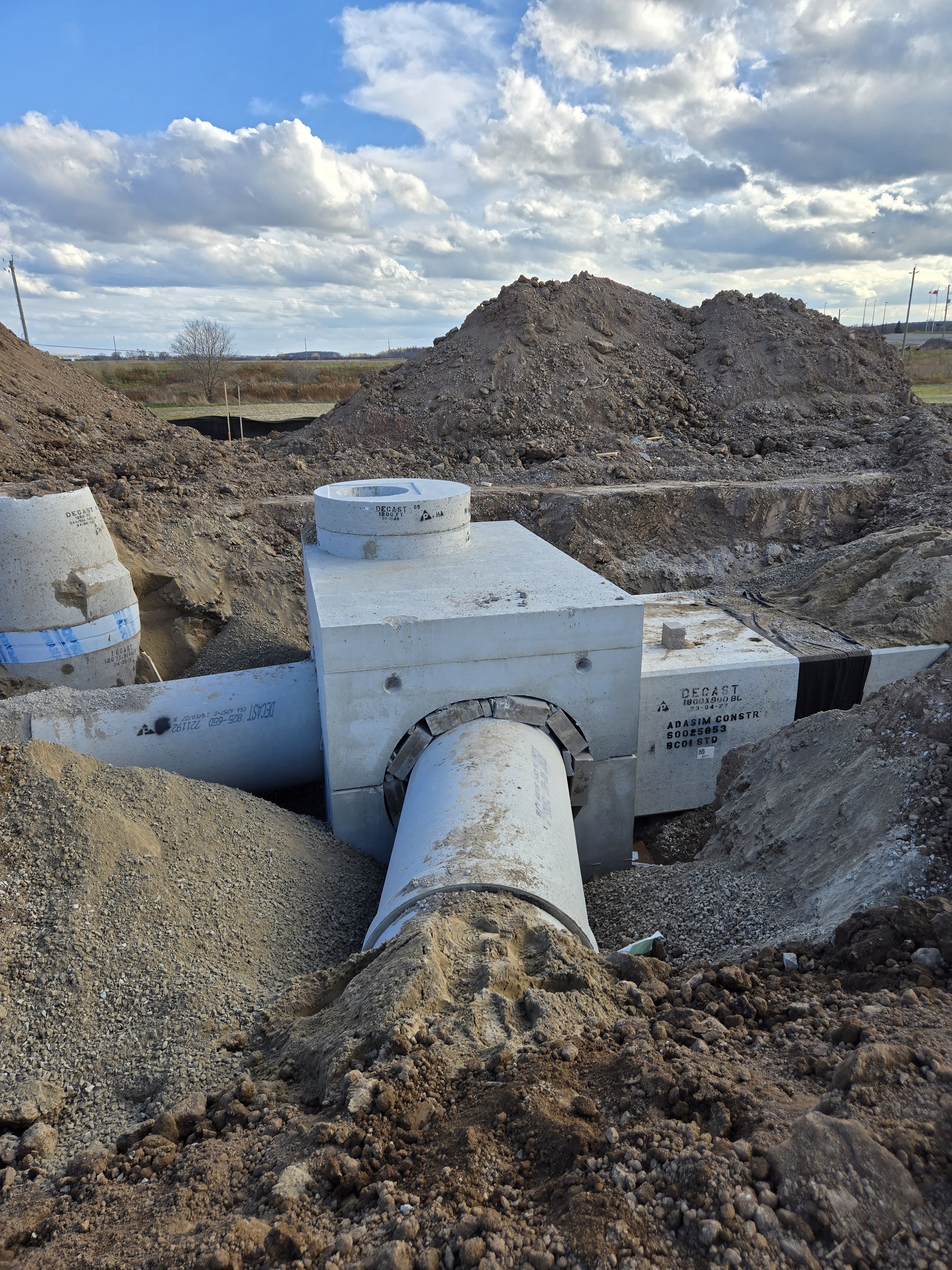 Construction site with large concrete pipes and dirt mounds under a partly cloudy sky.