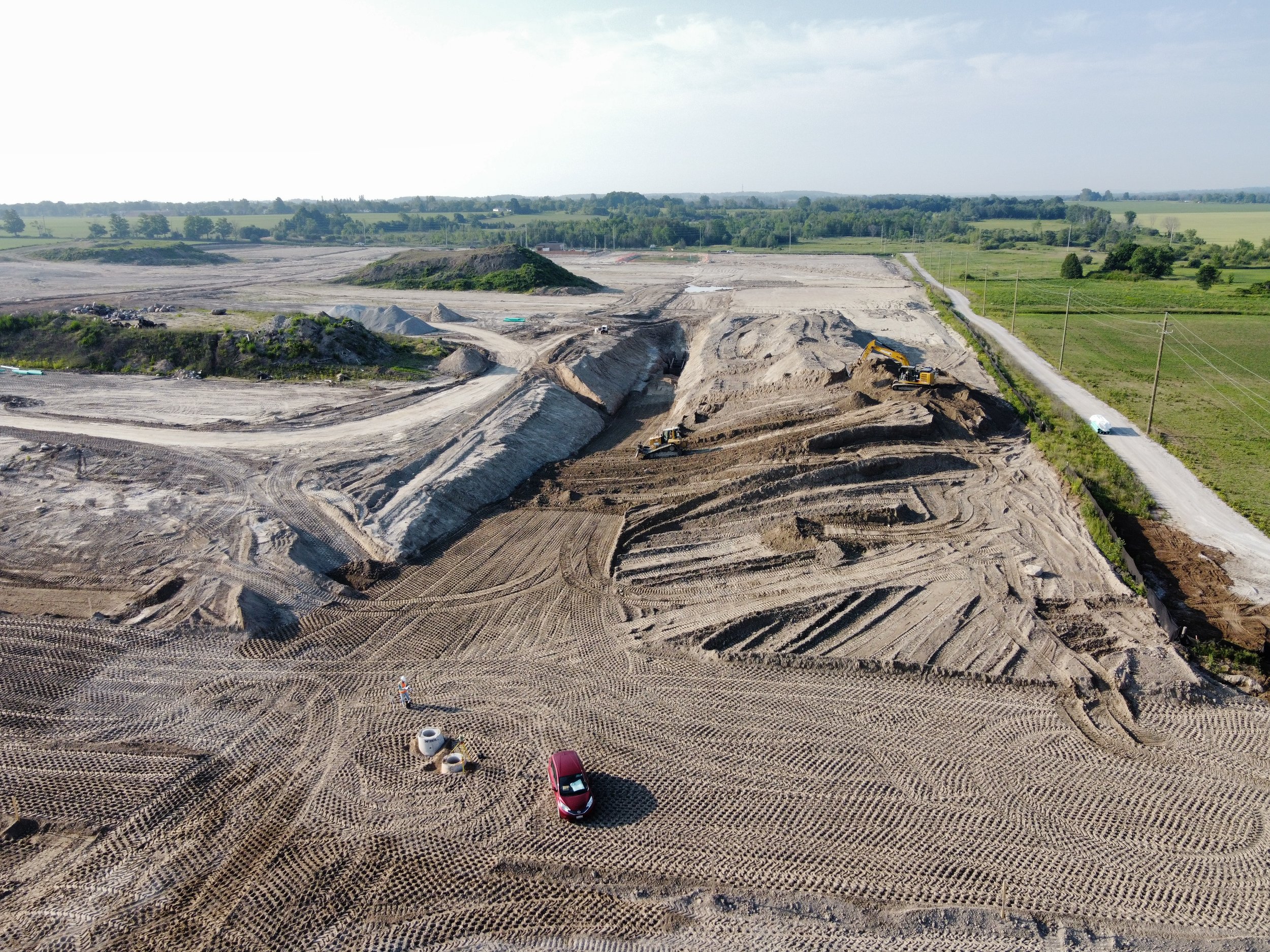 Aerial view of a construction site with earth-moving equipment, cleared land, and a dirt road connected to power poles, surrounded by green fields and trees in the background.