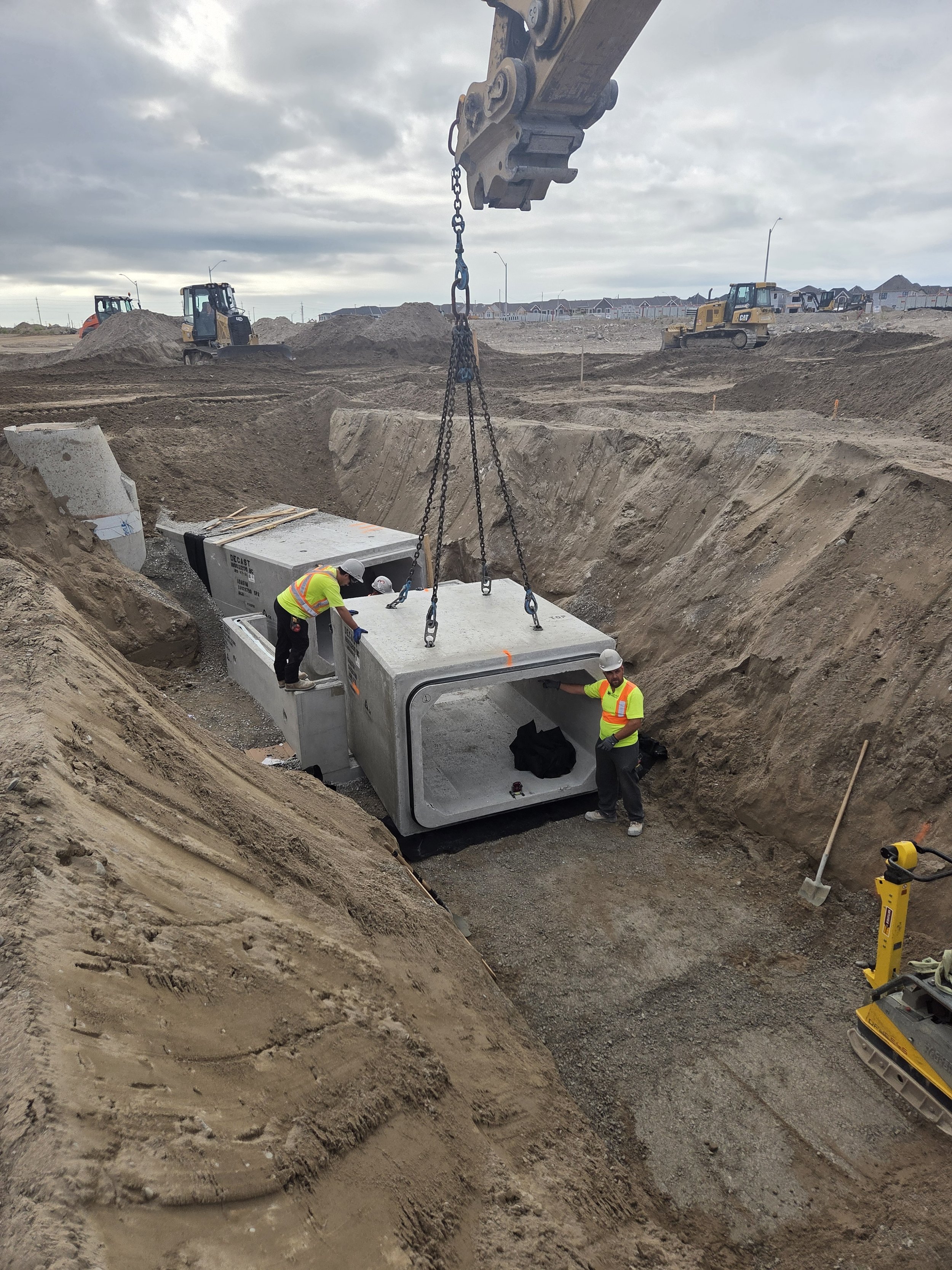 Construction workers install large concrete pipes in an excavated trench at a construction site with construction equipment and dirt mounds in the background.