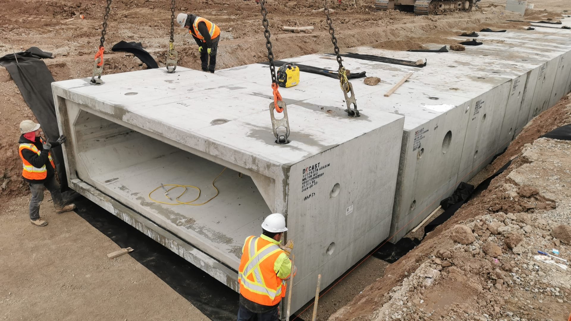 Construction workers in safety vests and helmets install a large precast concrete bridge section at a construction site.