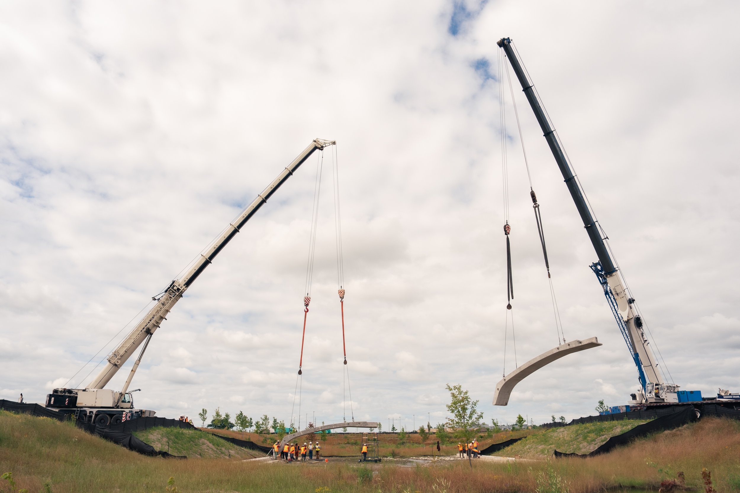 Construction site with two large cranes lifting concrete bridge segments over a grassy area, workers in safety vests and helmets on the ground, under a cloudy sky.