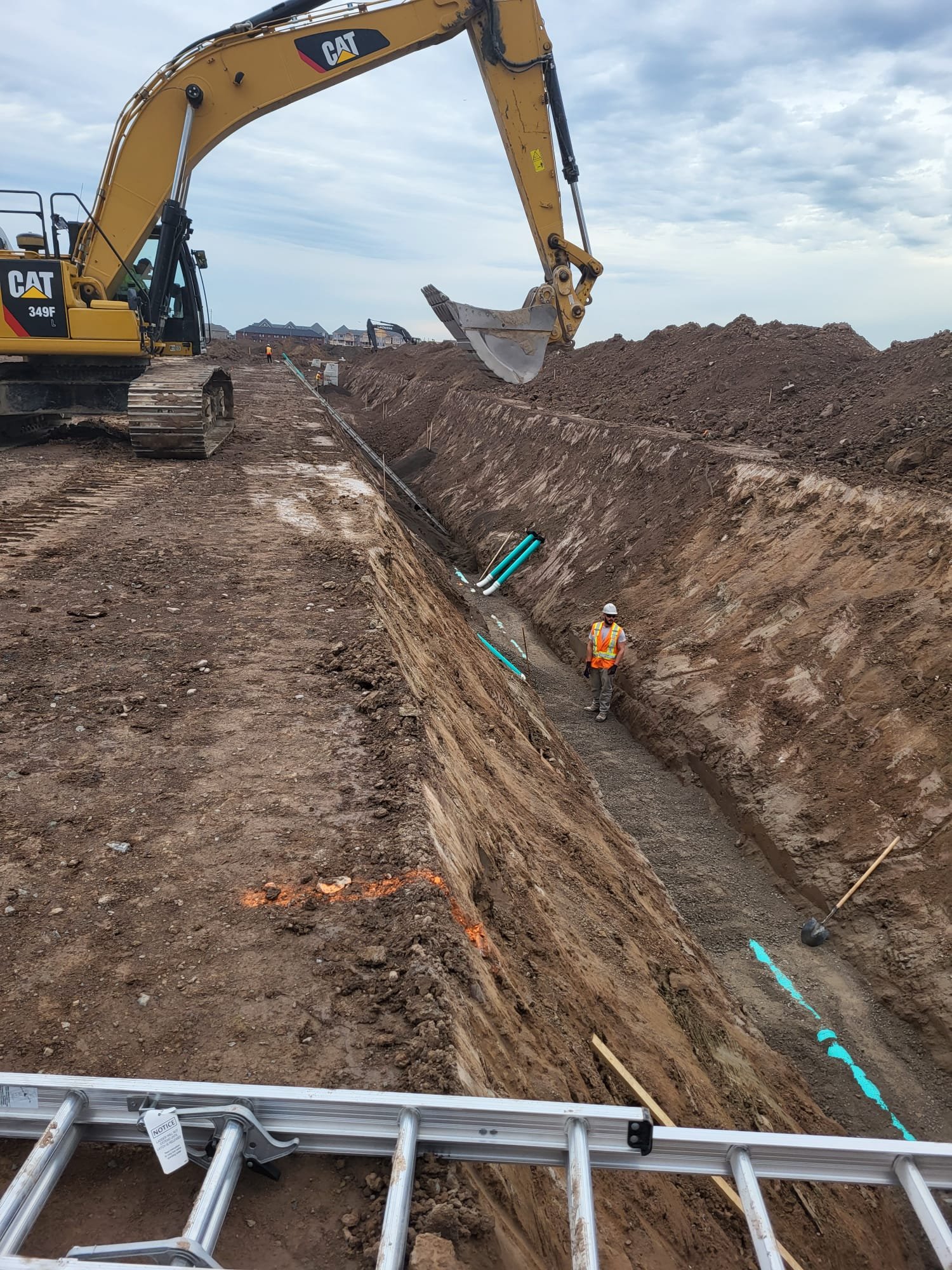 Construction site with a large yellow Caterpillar excavator on a dirt road digging a trench. Workers are inside the trench wearing safety gear. Blue pipes are being installed in the trench, and a metal ladder is in the foreground.