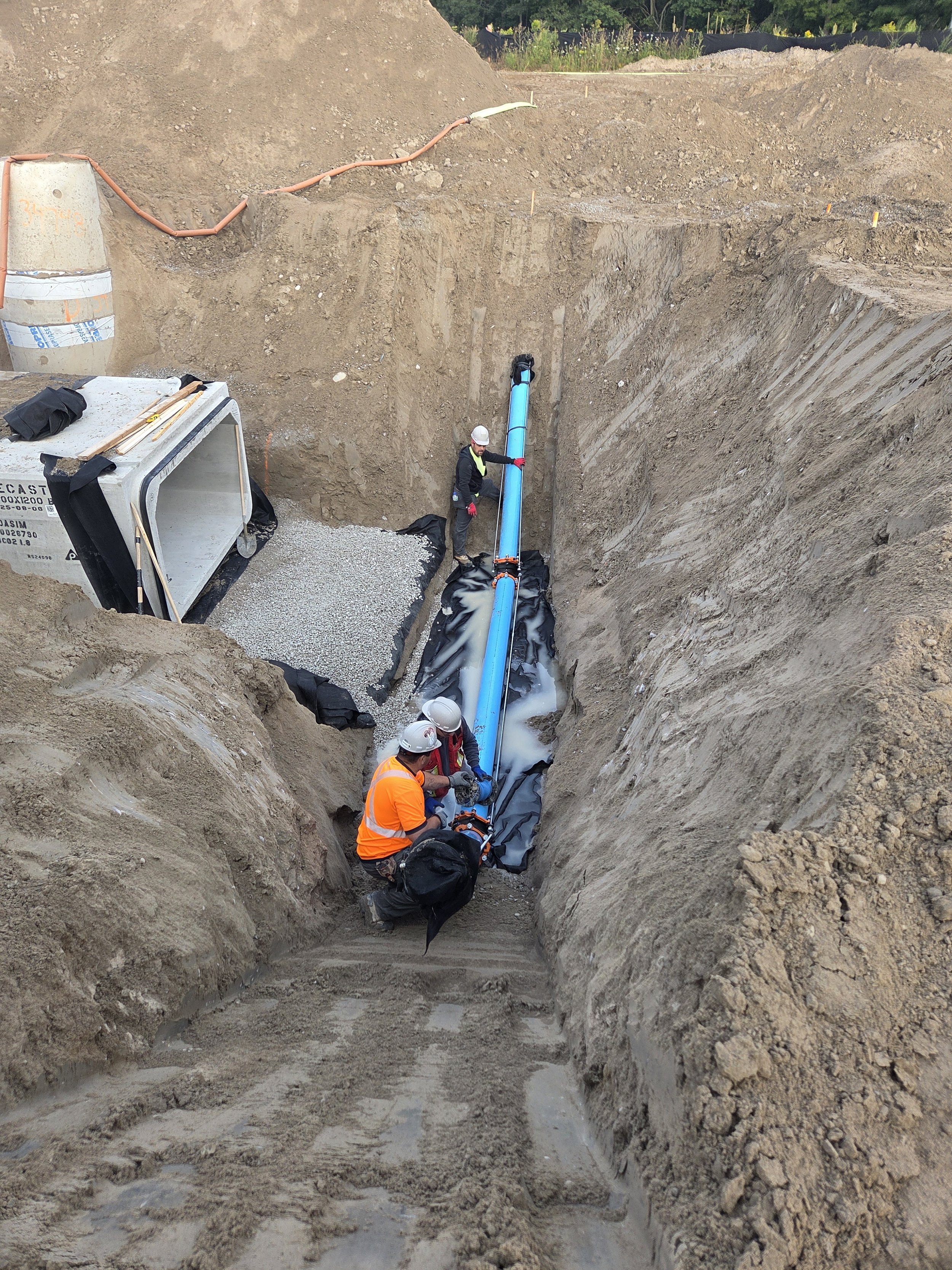 Construction workers installing a large blue pipe in an excavated trench at a construction site.