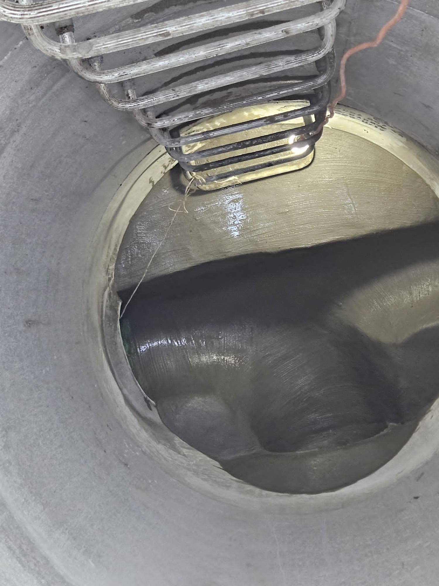 Inside of a concrete or metal cylindrical tank or pipe, showing a mixing or heating coil at the top and a black substance or material at the bottom.