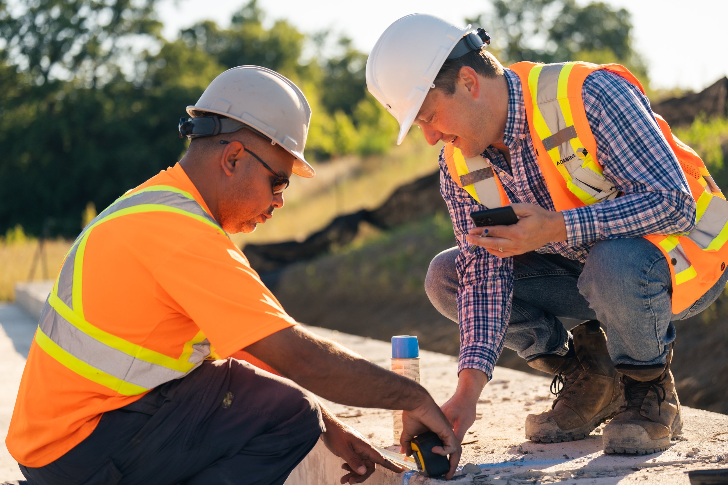 Two construction workers wearing helmets and safety vests working on a project outdoors, one kneeling and using a measuring tape while the other crouching and holding a phone, in a sunny environment with trees in the background.