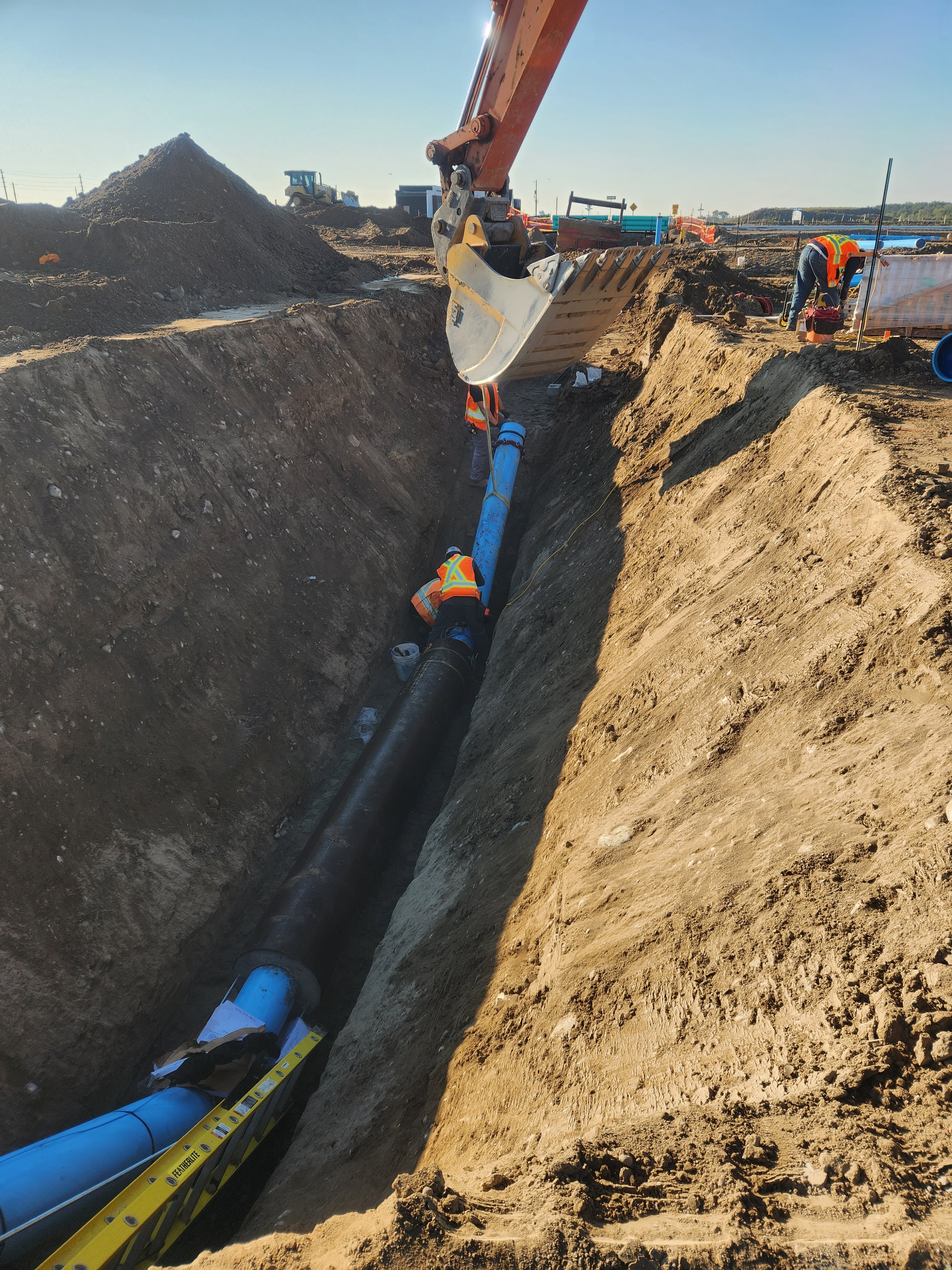 Workers installing large pipes in a deep construction trench at a construction site, with an excavator bucket overhead.