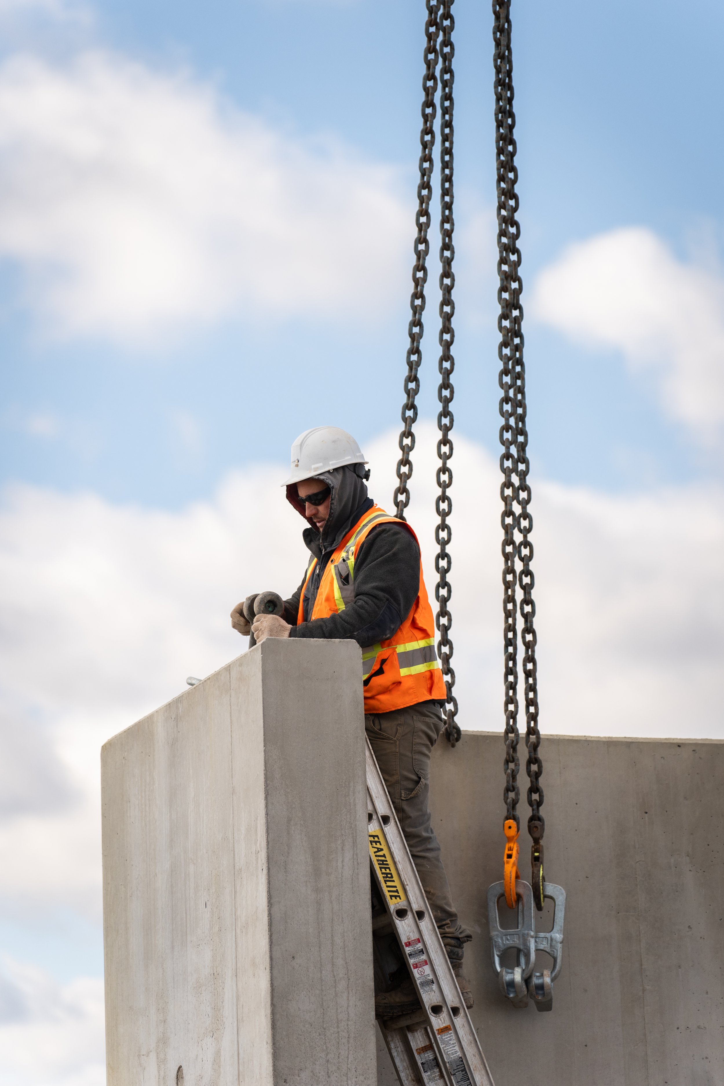 Construction worker wearing safety gear and a helmet fixing or inspecting a large concrete structure on a ladder outdoors.