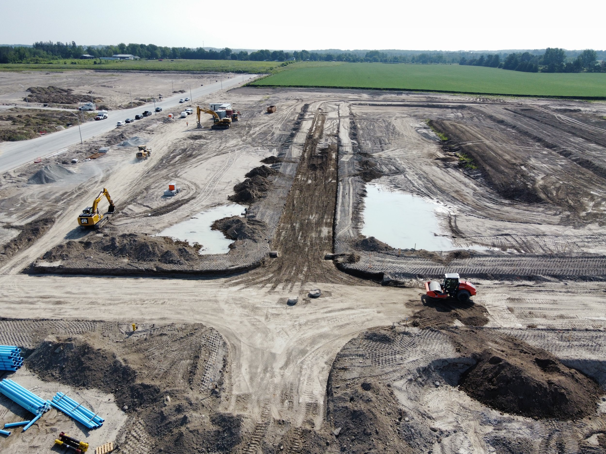 Construction site with excavators, trucks, and muddy areas, some with water, and construction materials, with an open field in the background.