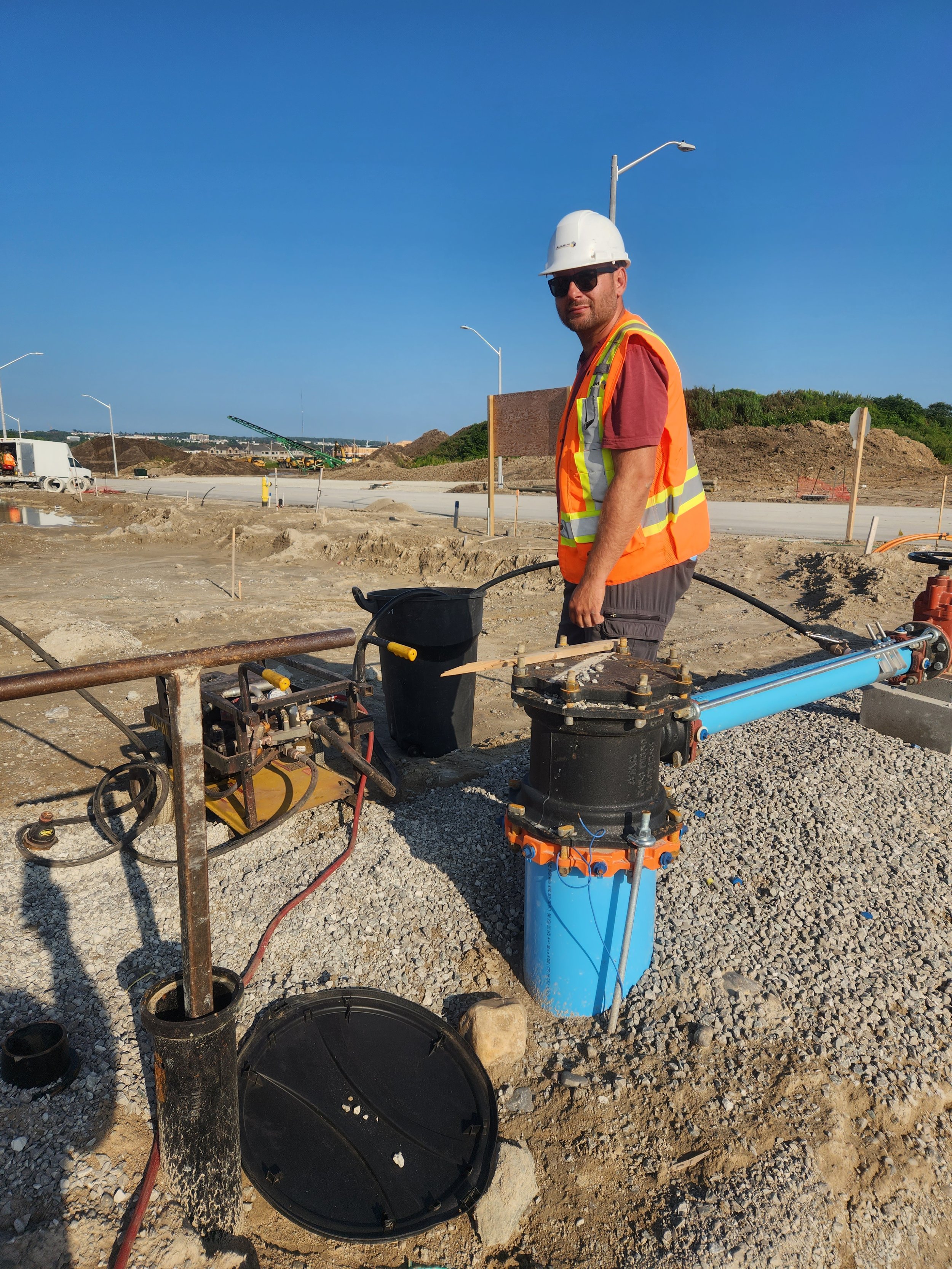 Construction worker wearing a white hard hat, sunglasses, an orange safety vest, and a maroon shirt standing next to construction machinery on a dirt site with gravel and piles of dirt, under a clear blue sky.
