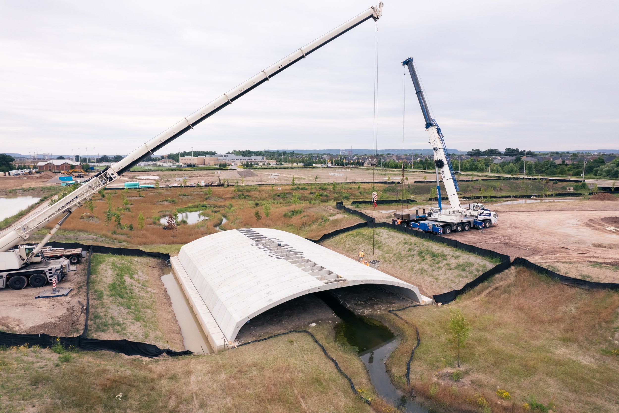 Construction site with two large cranes and a covered drainage tunnel, over a small stream, on overcast day.