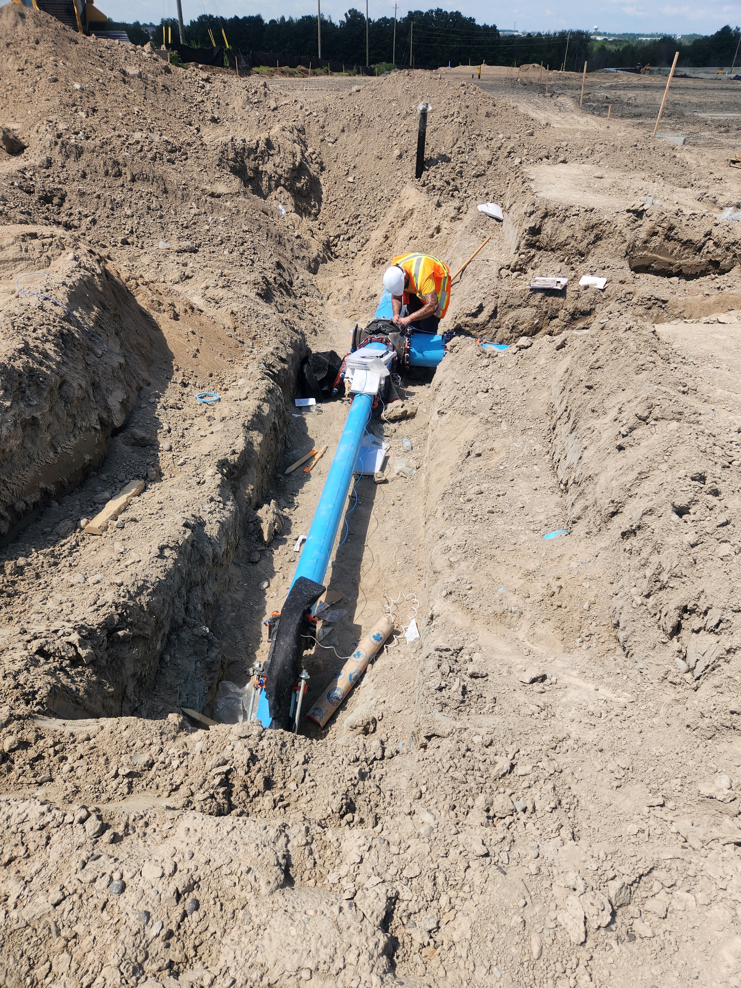 A construction worker in a yellow safety vest and white helmet working in a trench, installing or maintaining large blue pipes at a construction site with exposed dirt and equipment.