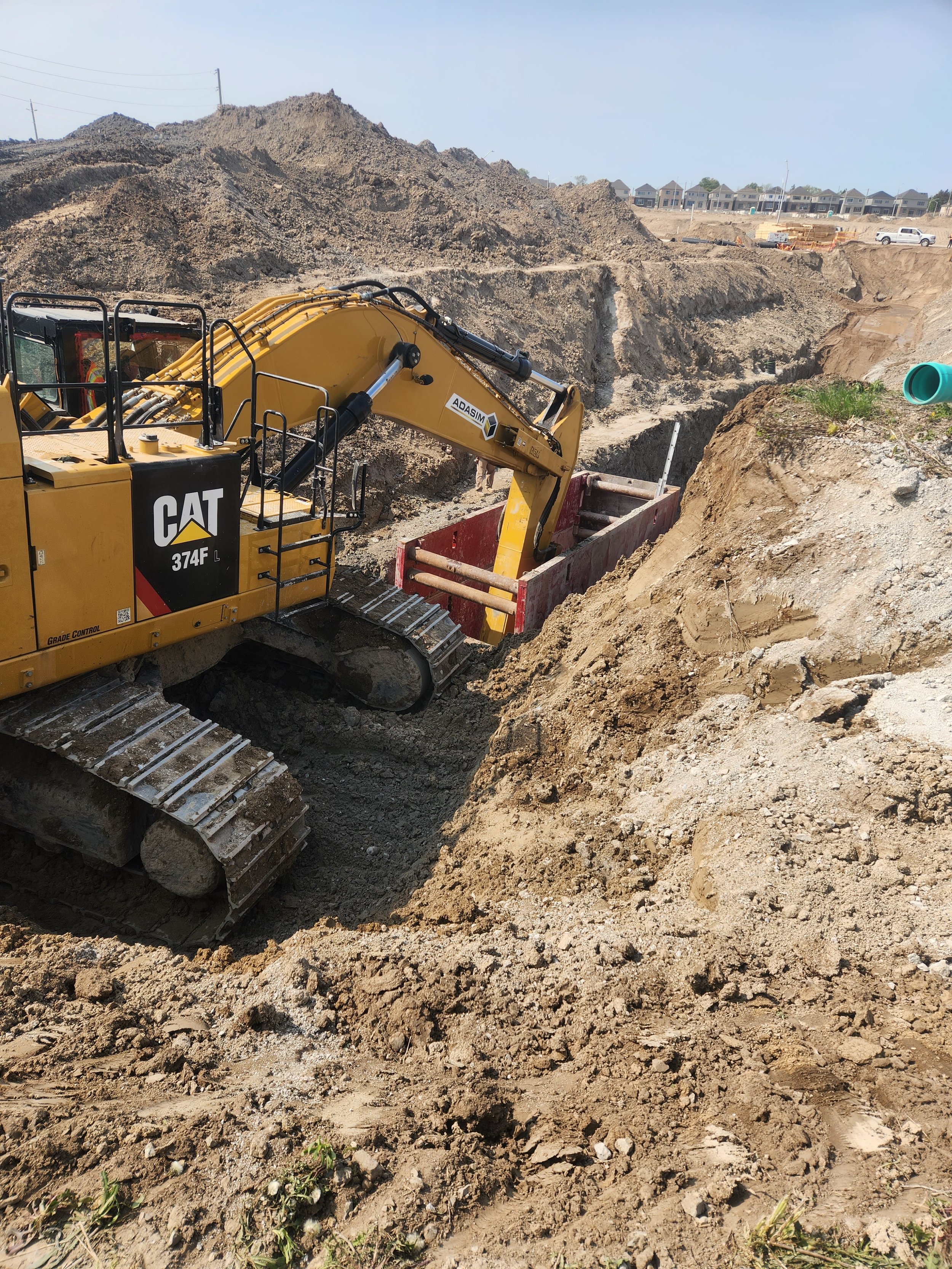 A yellow CAT 374F excavator working in a construction site, digging into the earth and moving soil with a large red trench box. In the background, there are dirt mounds, construction partial structures, and houses under a clear sky.