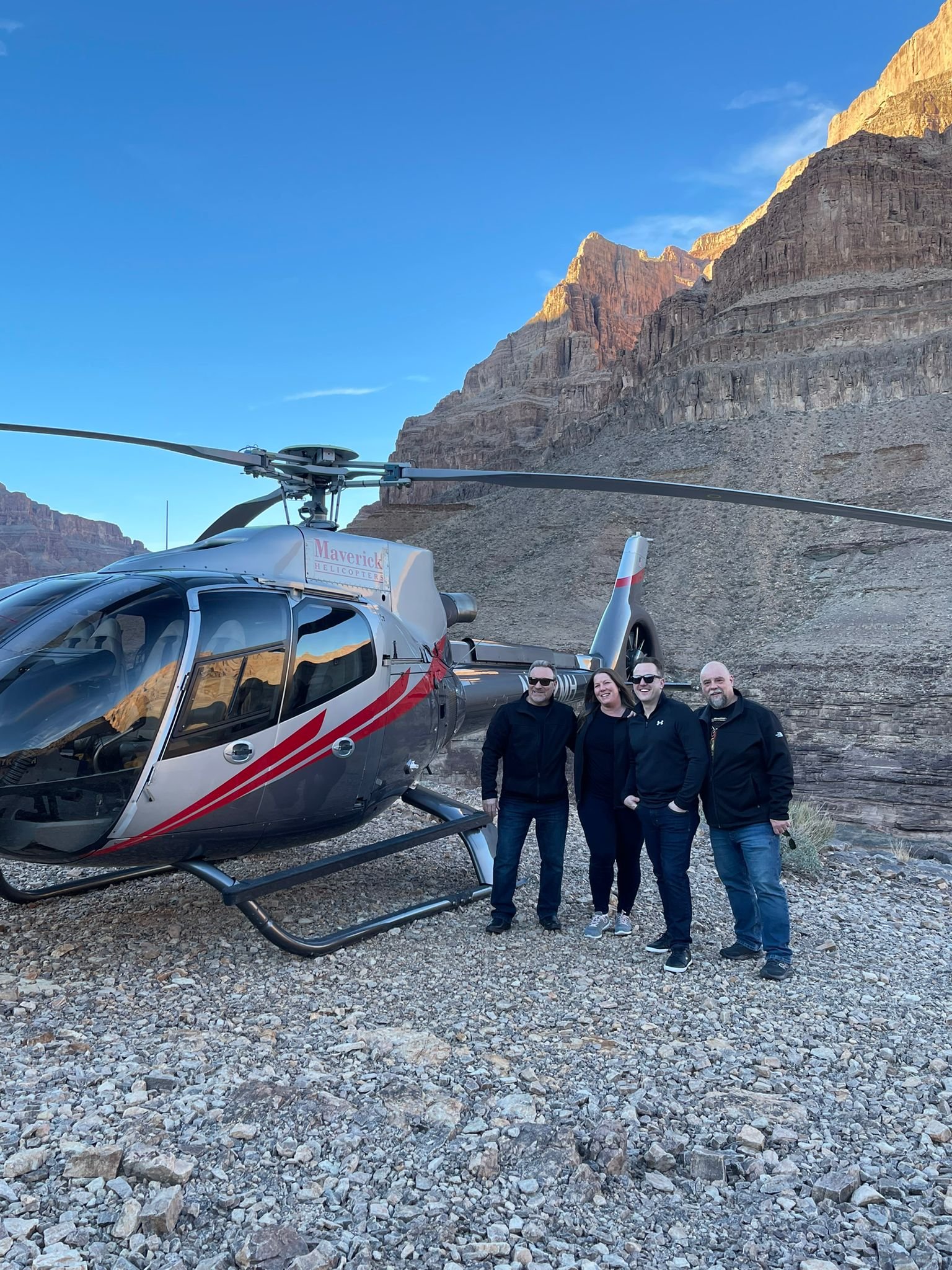 Four adults standing next to a helicopter at the Grand Canyon, with rocky terrain and canyon cliffs in the background.