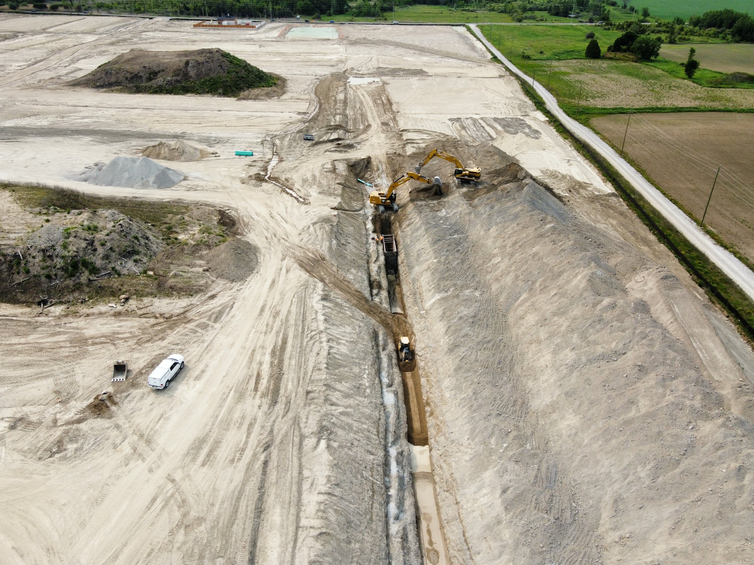 Construction site with excavators digging a trench, dirt piles, and construction vehicles. Open land and fields are visible in the background.