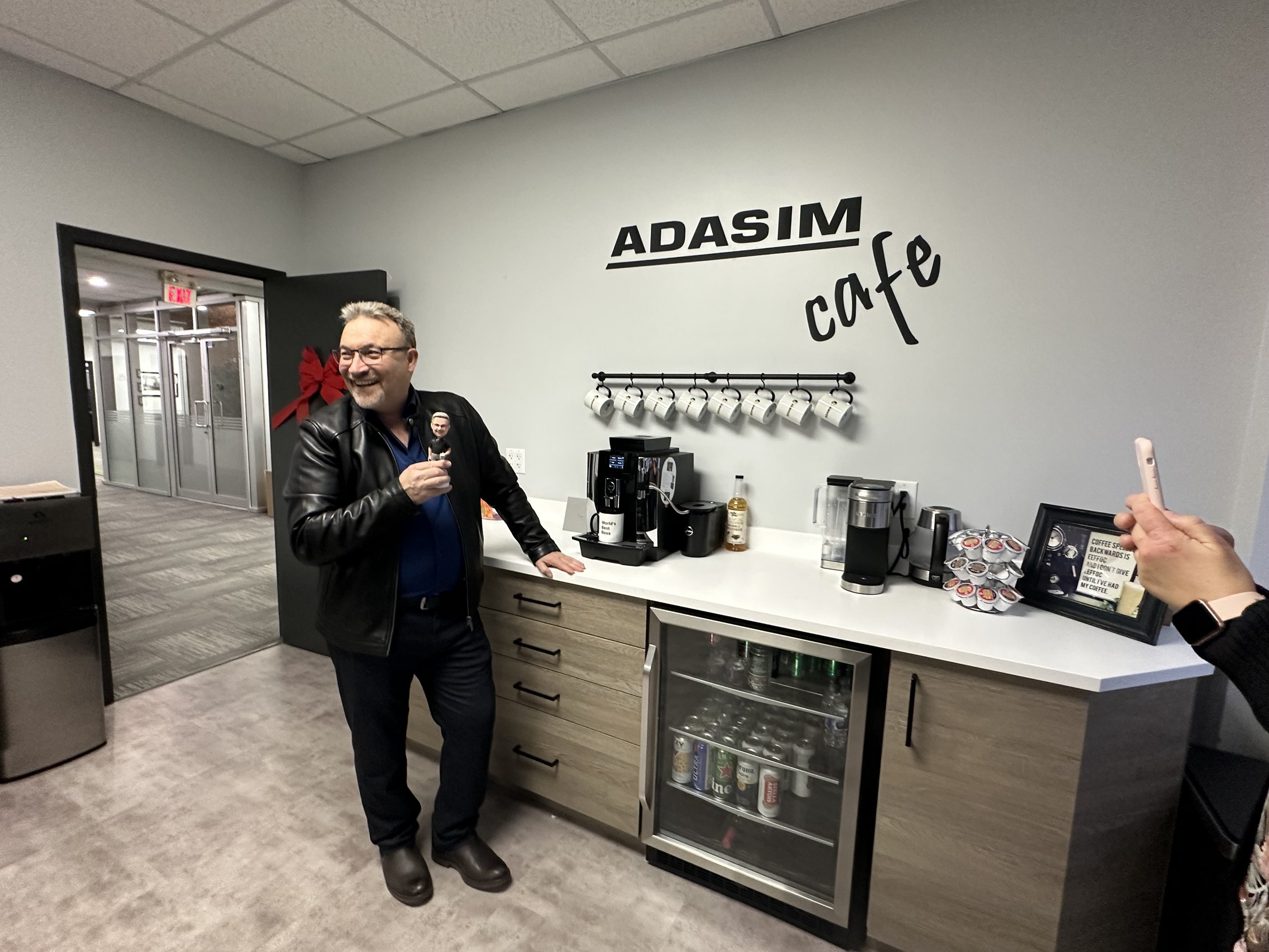 Man in black leather jacket and glasses standing in front of a coffee station at ADASIM cafe, smiling and holding a small object. Coffee cups hanging on a rack, coffee machines, and beverages in a glass-front fridge are visible behind the counter.