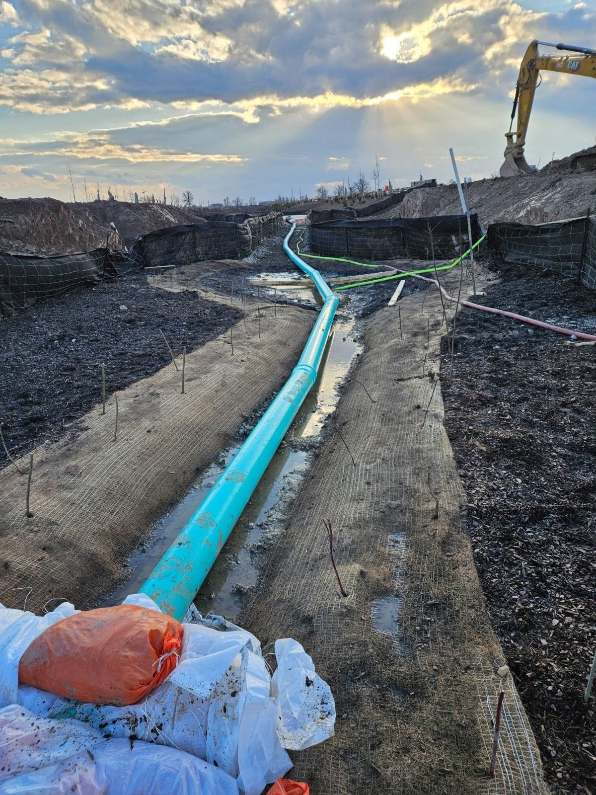 Construction site with an orange hose, blue pipe, and excavator in the background under a cloudy sky.