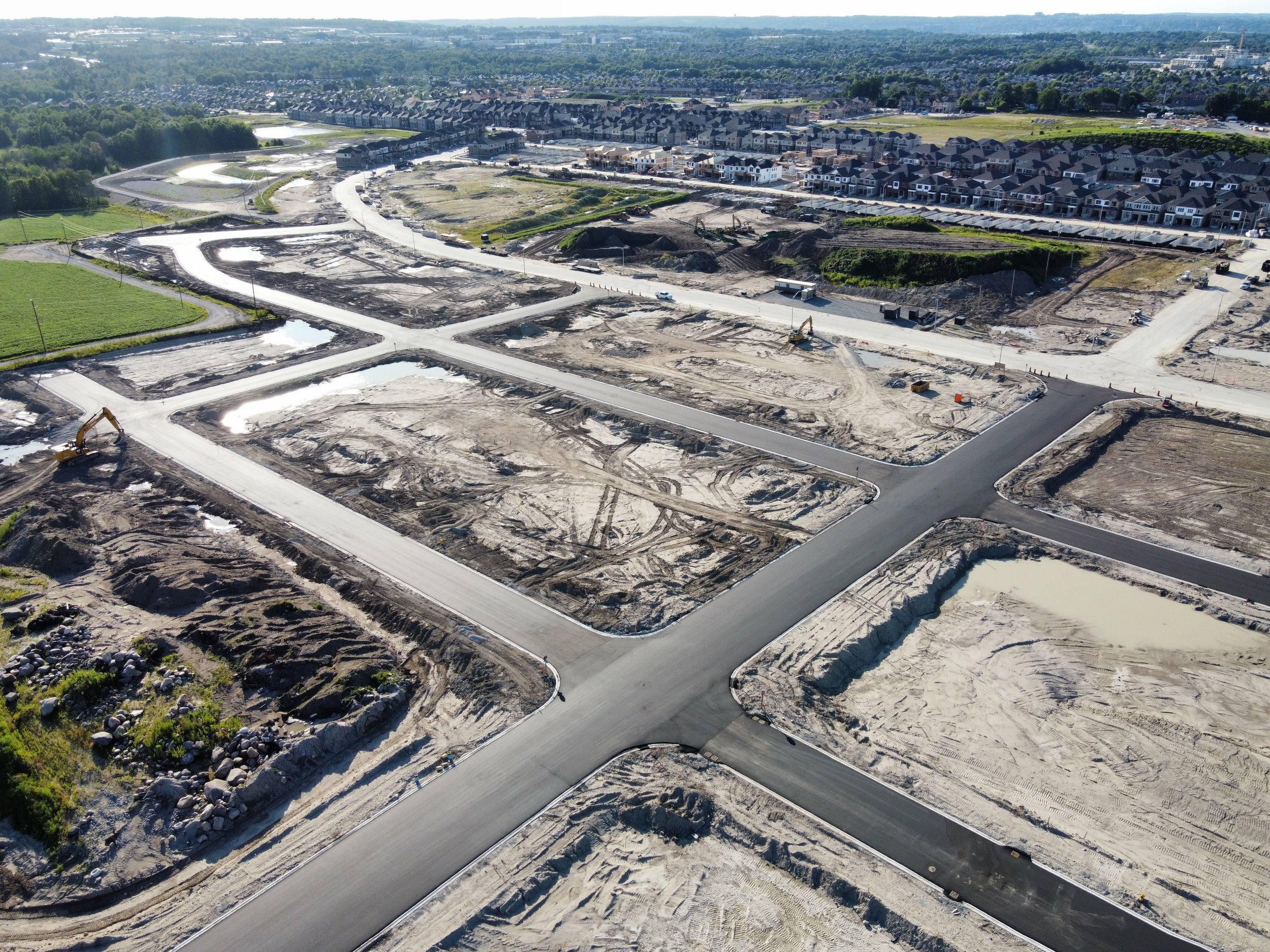 An aerial view of a construction site with roads being built, several excavators, and cleared plots of land, with a residential neighborhood in the background.