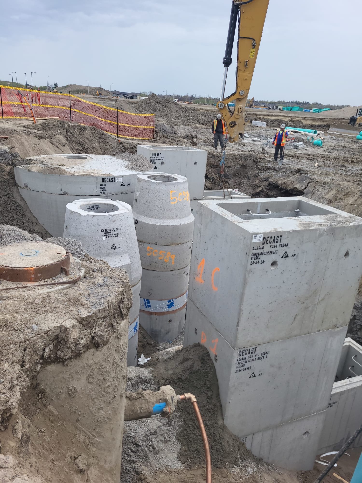 Construction site with large concrete drainage structures and sewer pipes being installed. Workers in safety vests and hard hats present, with construction equipment visible in the background.