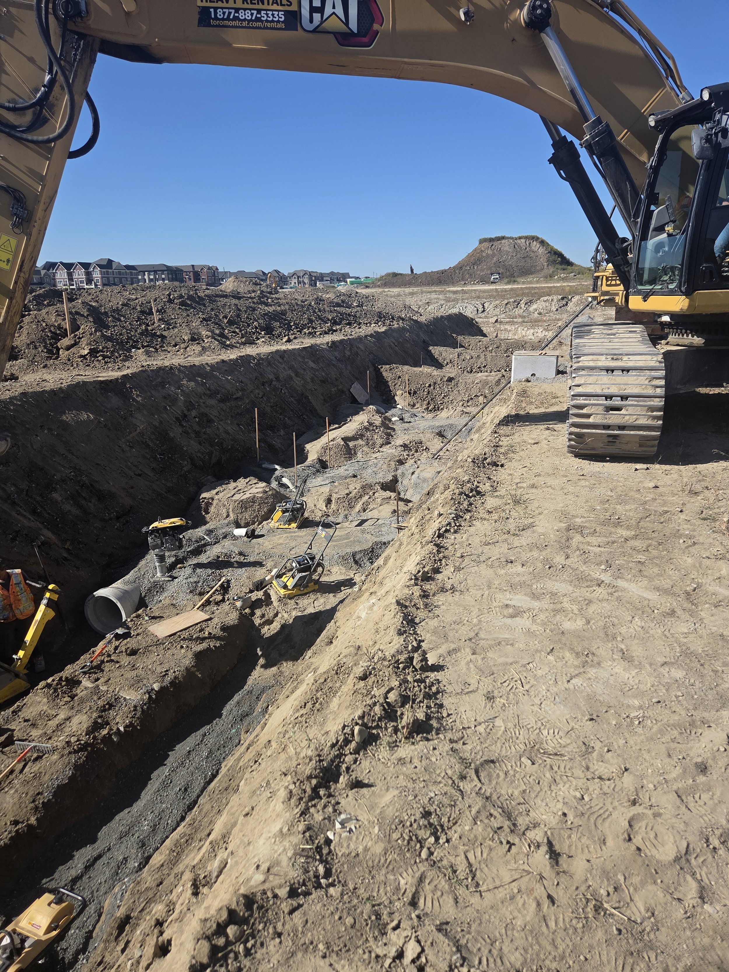 Construction site with excavator and digging trench for pipes, under clear blue sky in a developing area.