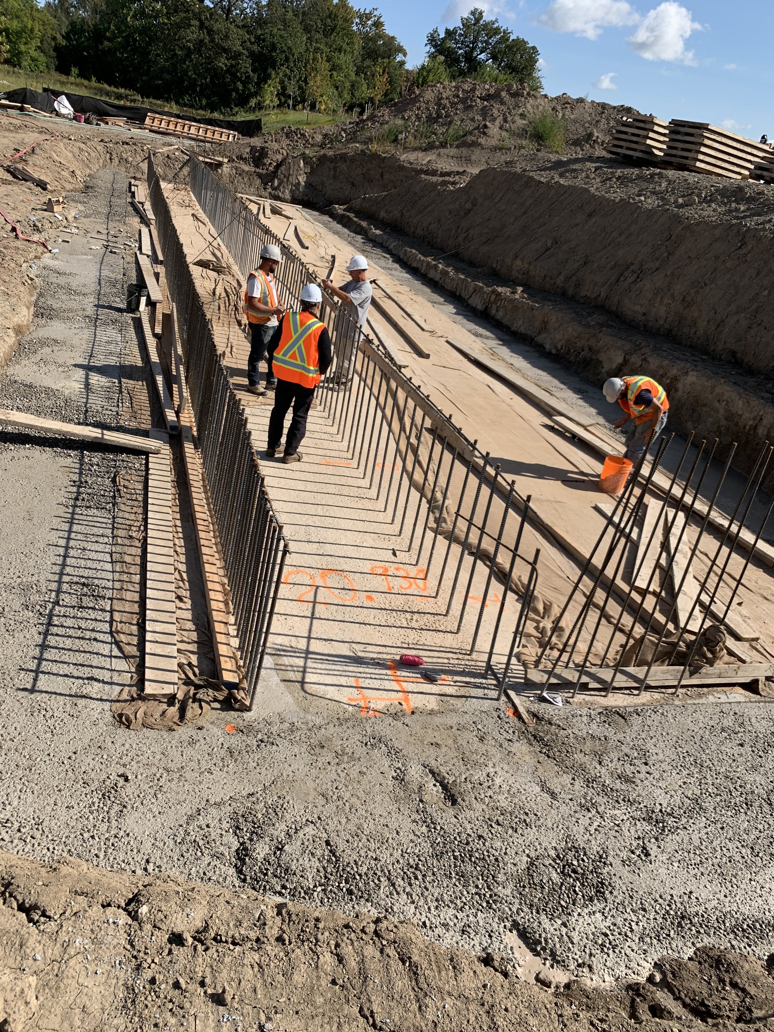 Construction workers wearing safety vests and helmets working on a building foundation, installing steel rebar and pouring concrete at a construction site.
