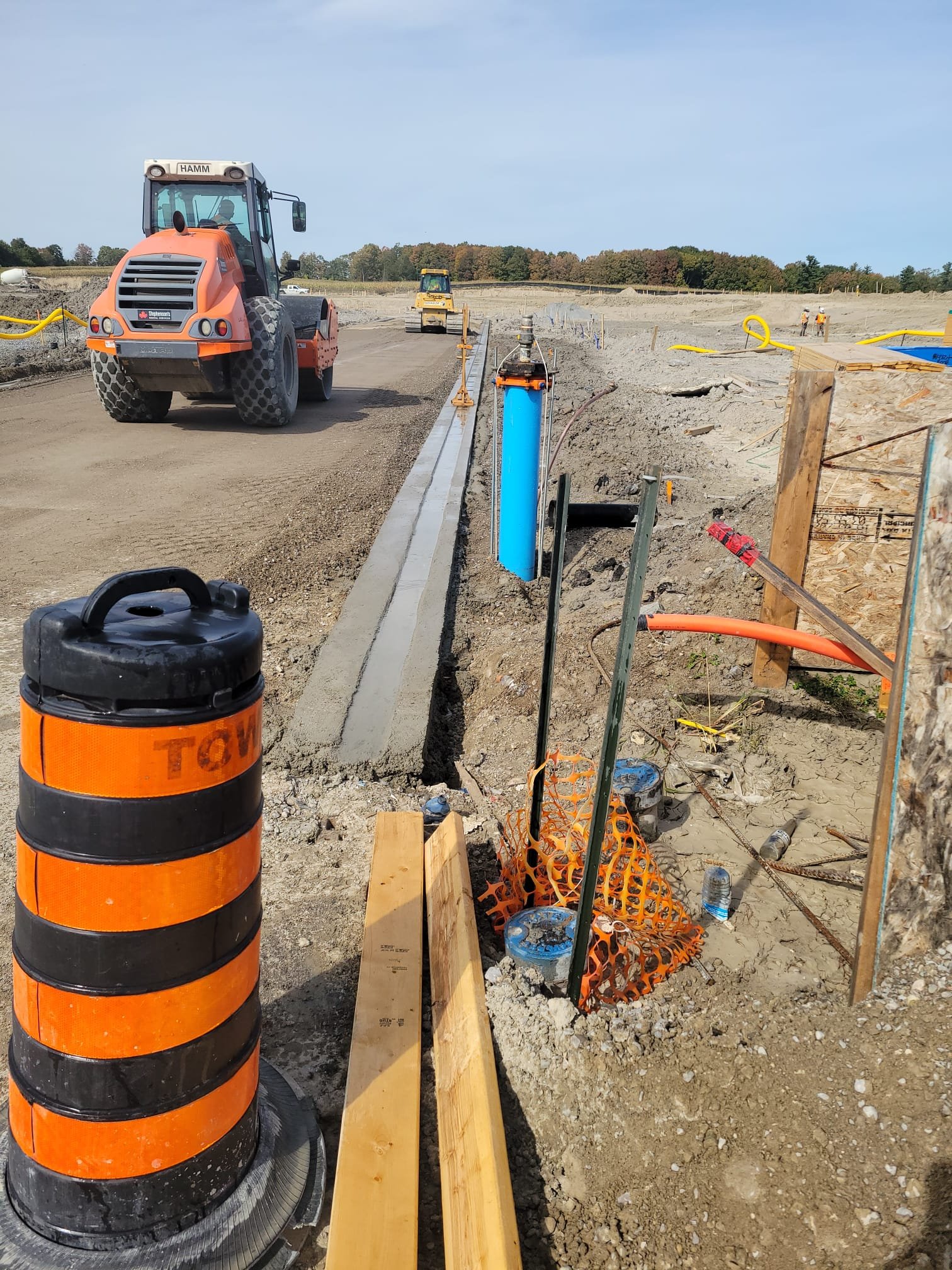 Construction site with heavy machinery, dirt road, and construction materials, including orange and black traffic barrels, wooden planks, and a blue utility pipe.