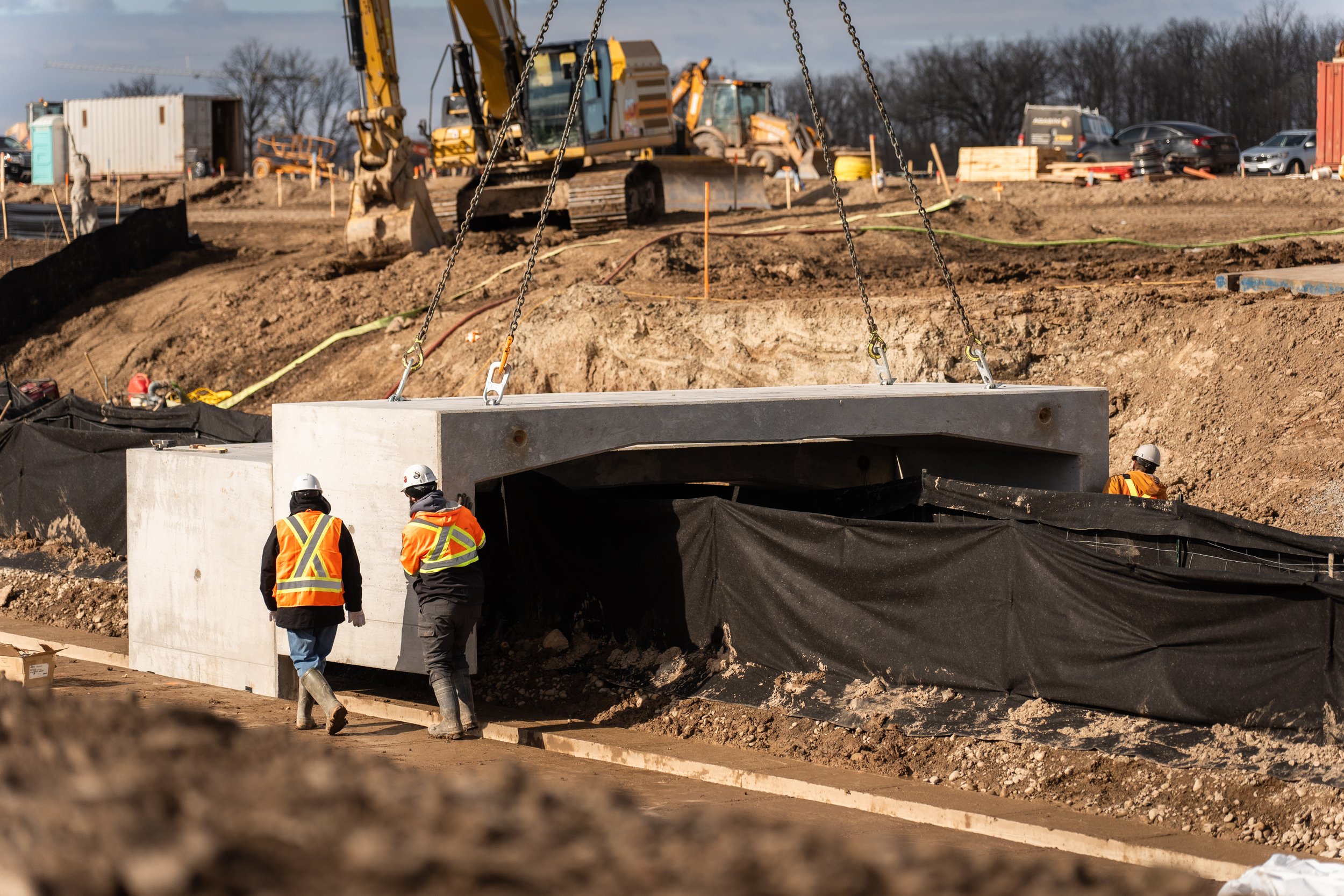 Construction workers install a large concrete tunnel segment at a construction site with excavators, dirt, and construction vehicles in the background.