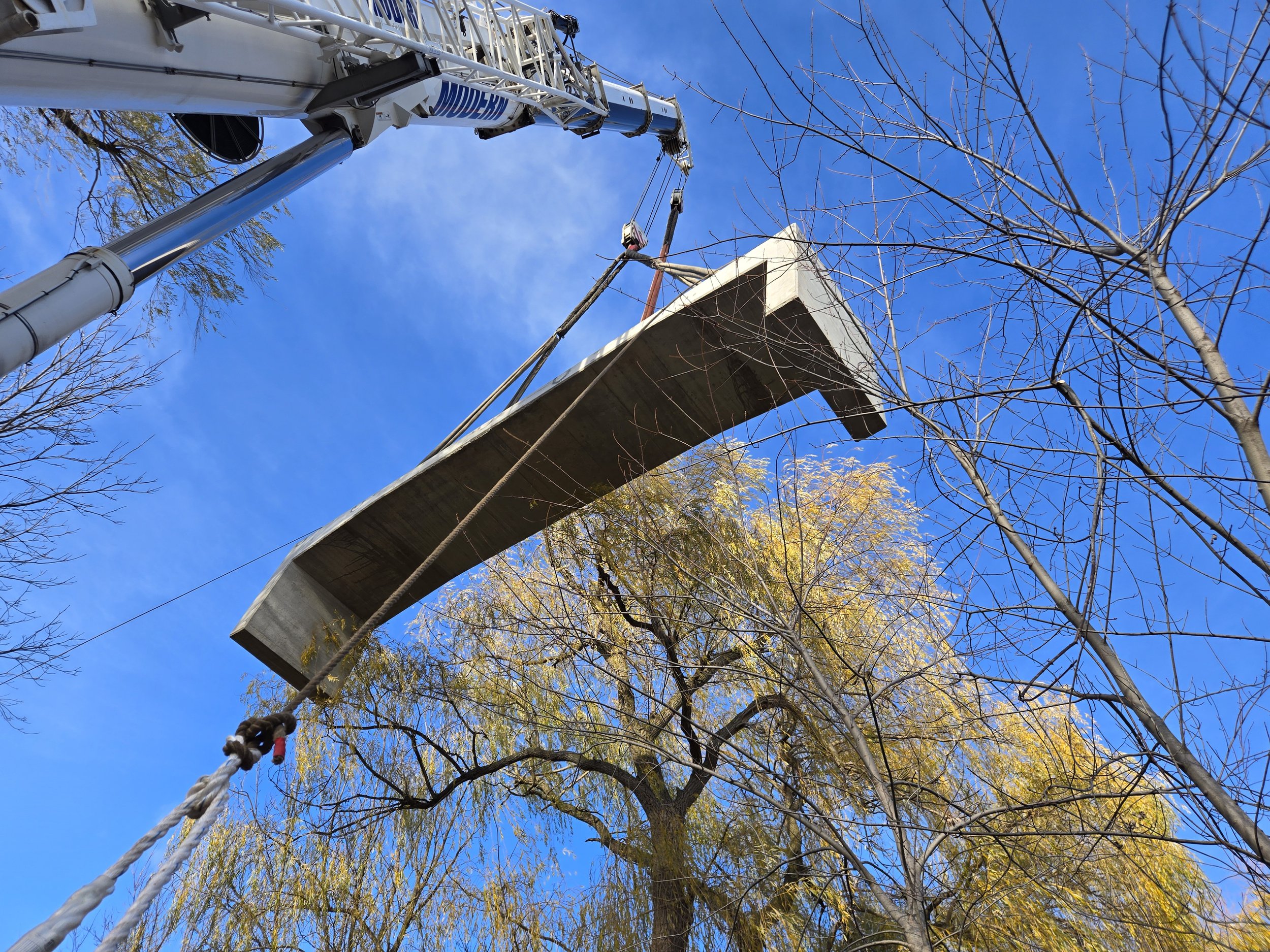A construction crane lifting a large concrete slab in a park with tall trees and a bright blue sky with some clouds.