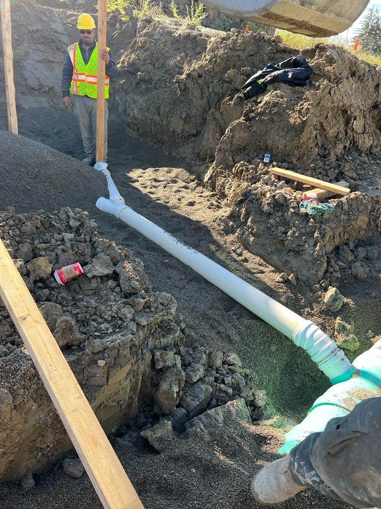 A construction worker wearing a yellow hard hat, sunglasses, and a fluorescent safety vest stands next to a long white pipe laid on the ground in a dirt excavation site. The site has rocky soil and a large open trench, with some construction material