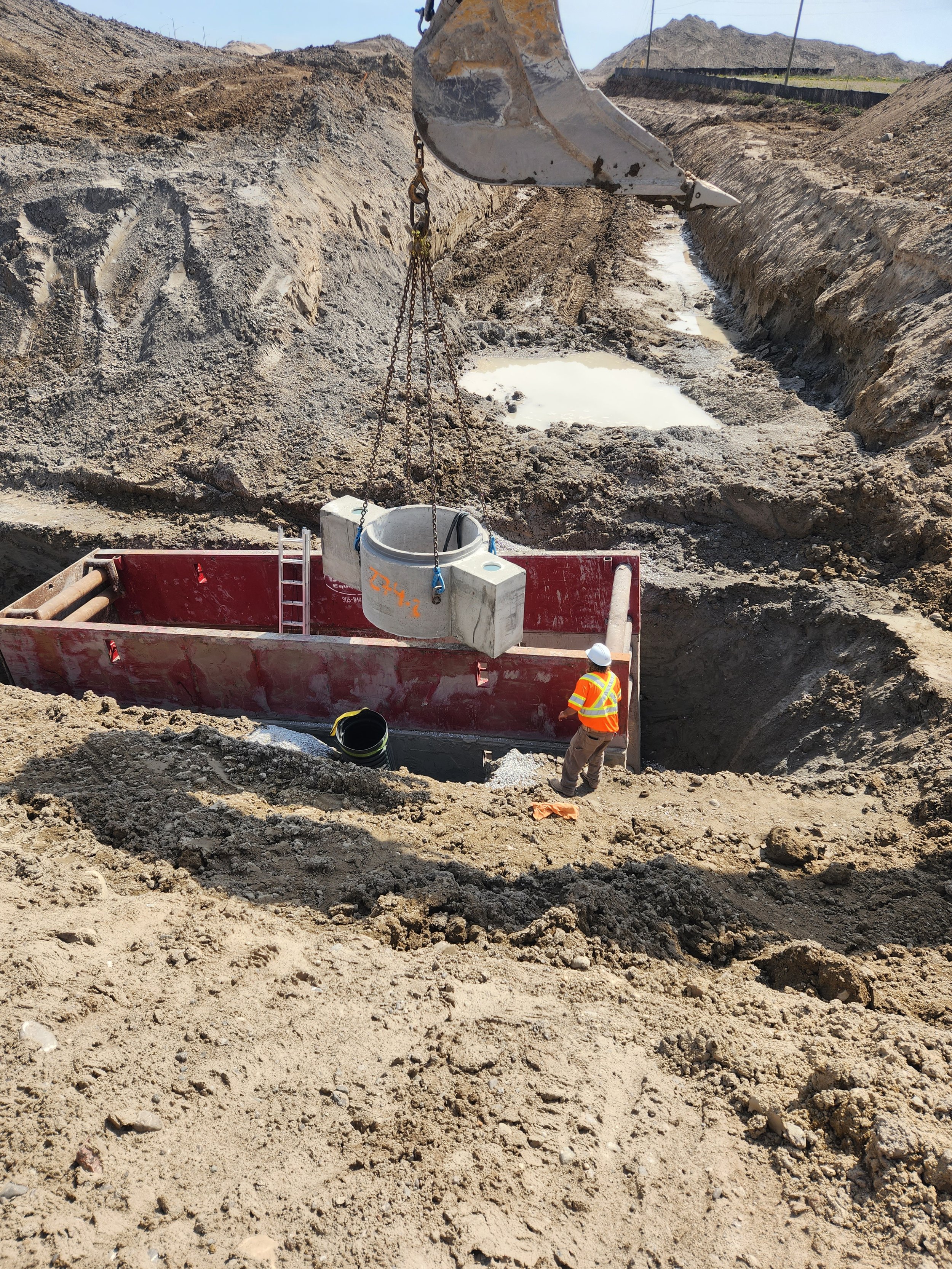 Construction worker in safety vest and helmet standing in trench on a construction site, with a crane lifting a concrete pipe and a red barrier surrounding the work area, landscape of dirt mounds and rocks in the background.
