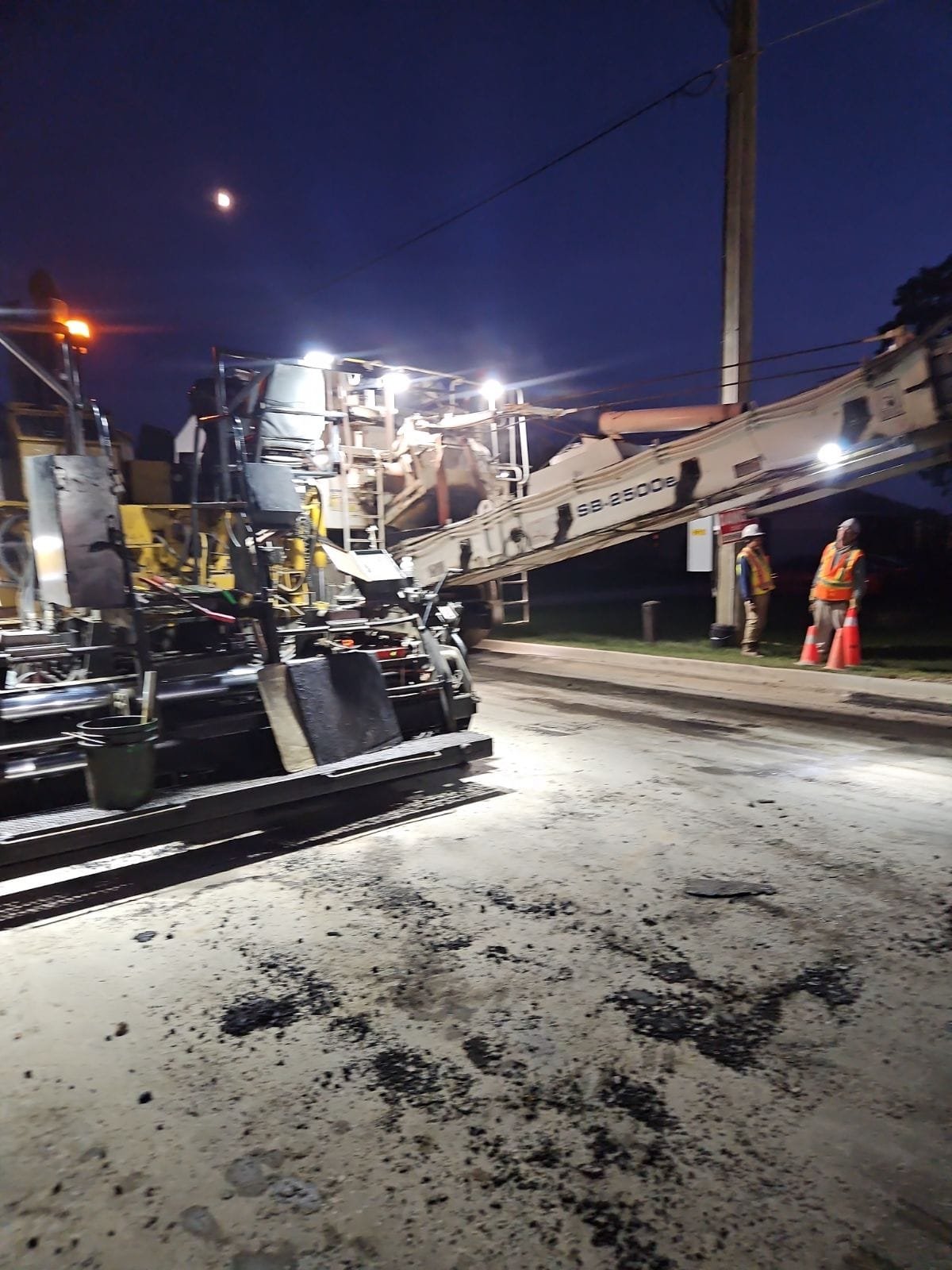 Construction workers wearing safety vests and helmets working near a large road construction machine on a street at night, with a clear dark sky and a visible moon.