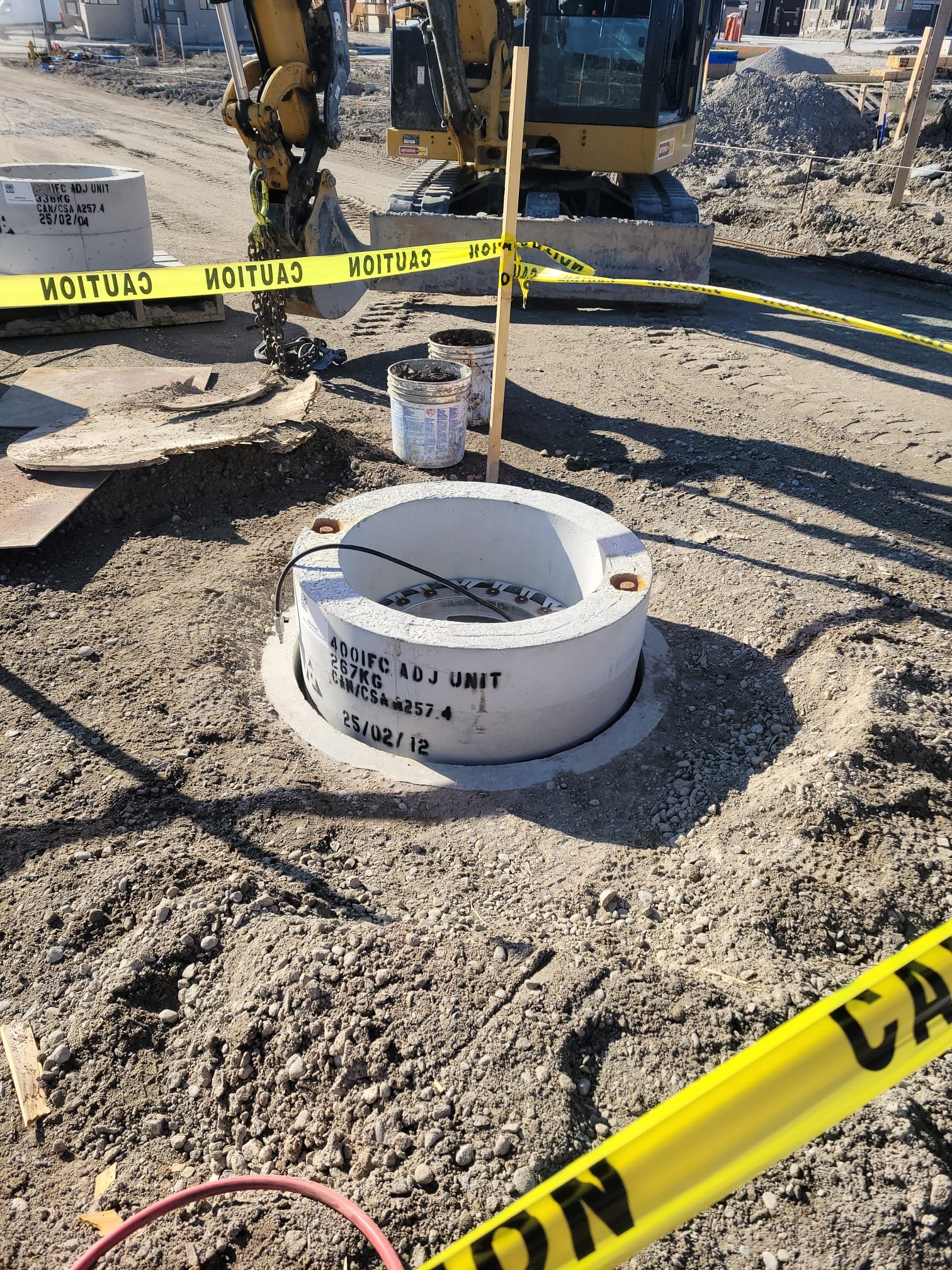Construction site with a large valve or pipe fitting labeled '400IEC ADJ UNIT 27KG' surrounded by caution tape, with a backhoe, tools, and construction materials in the background.
