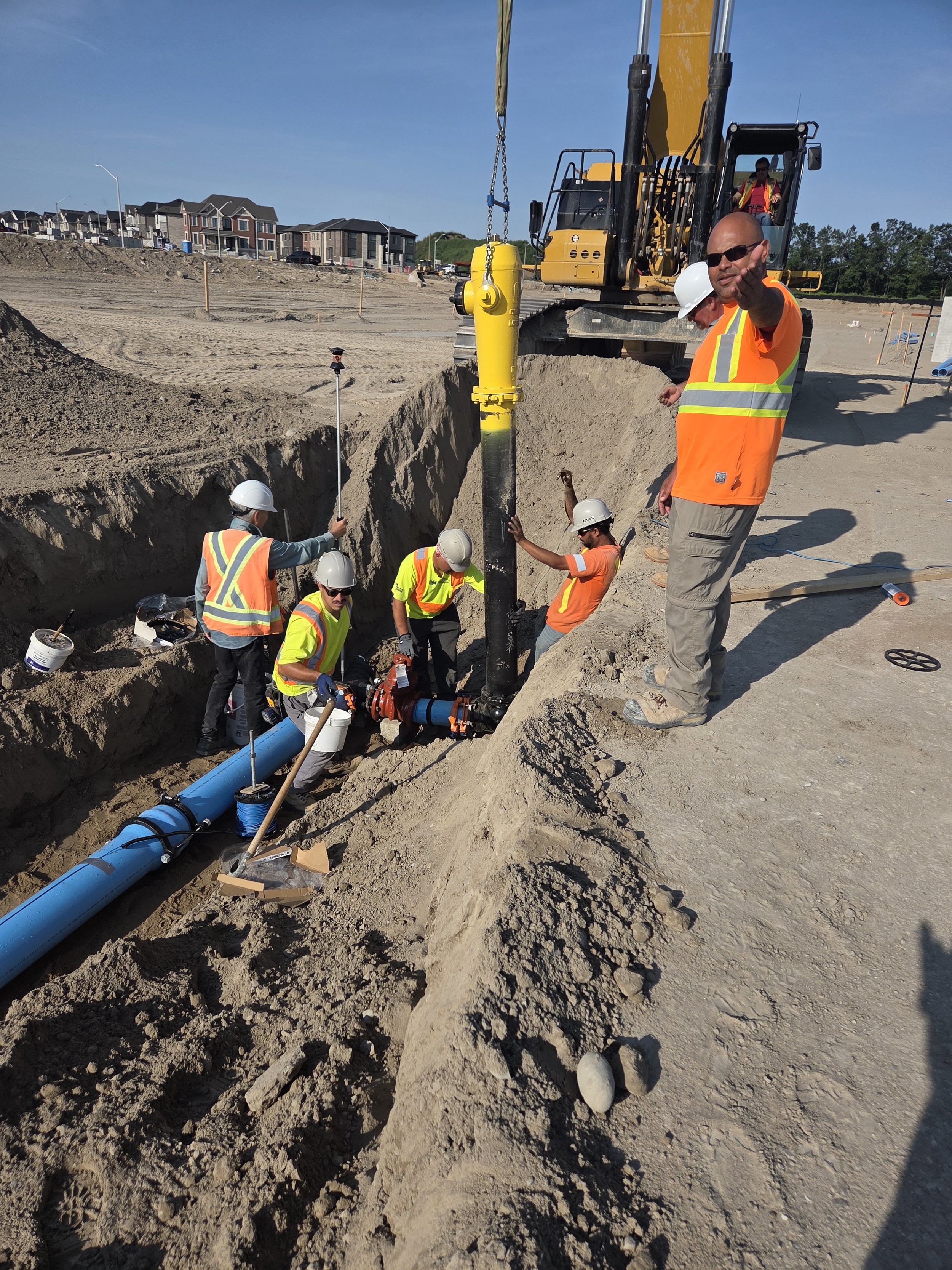 Construction workers installing a large black pipe underground, with a crane lifting a yellow pipe fitting, at a construction site with residential buildings in the background.