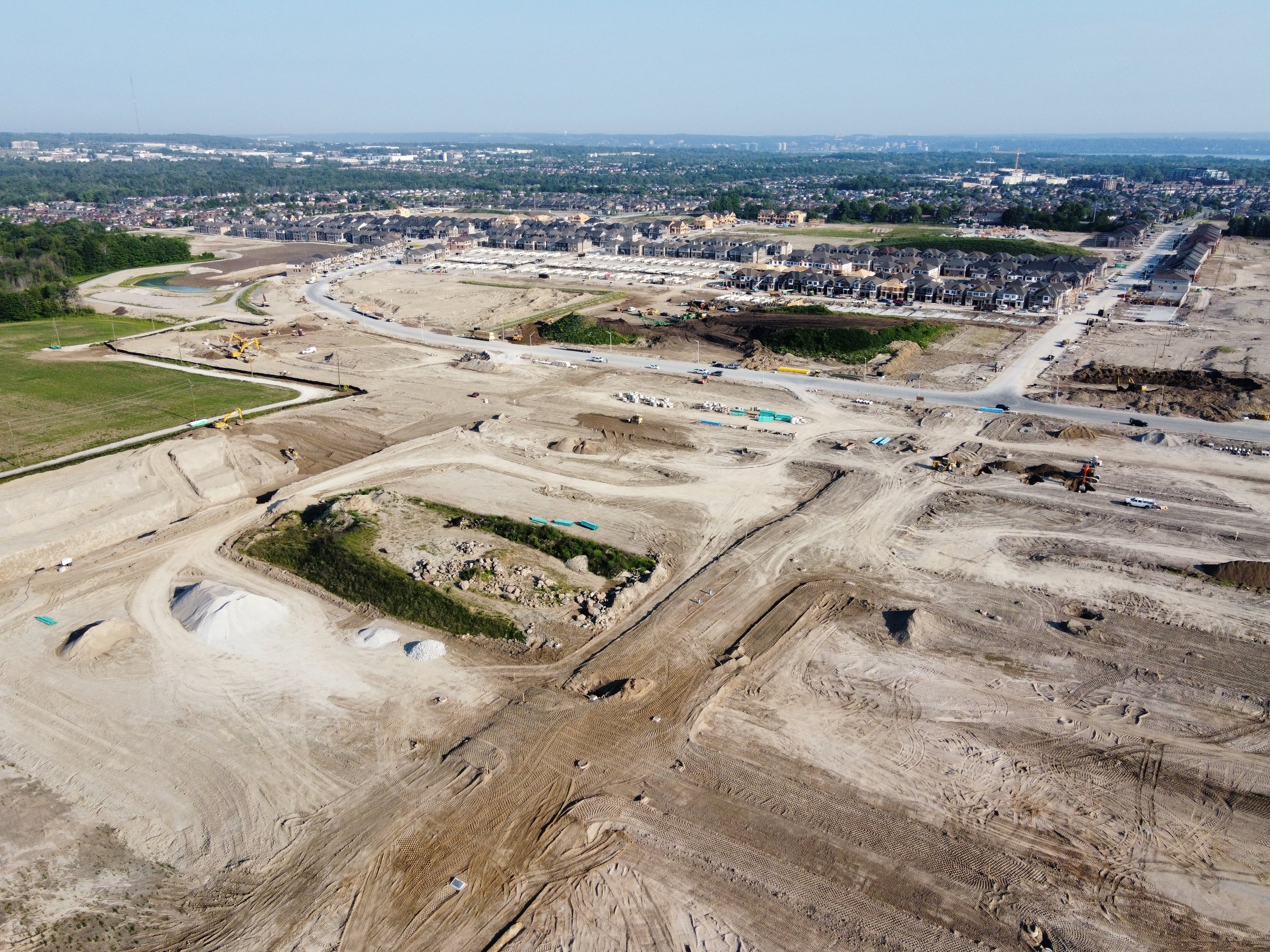 An aerial view of a construction site with dirt roads, excavations, and construction vehicles, with a residential neighborhood in the background.