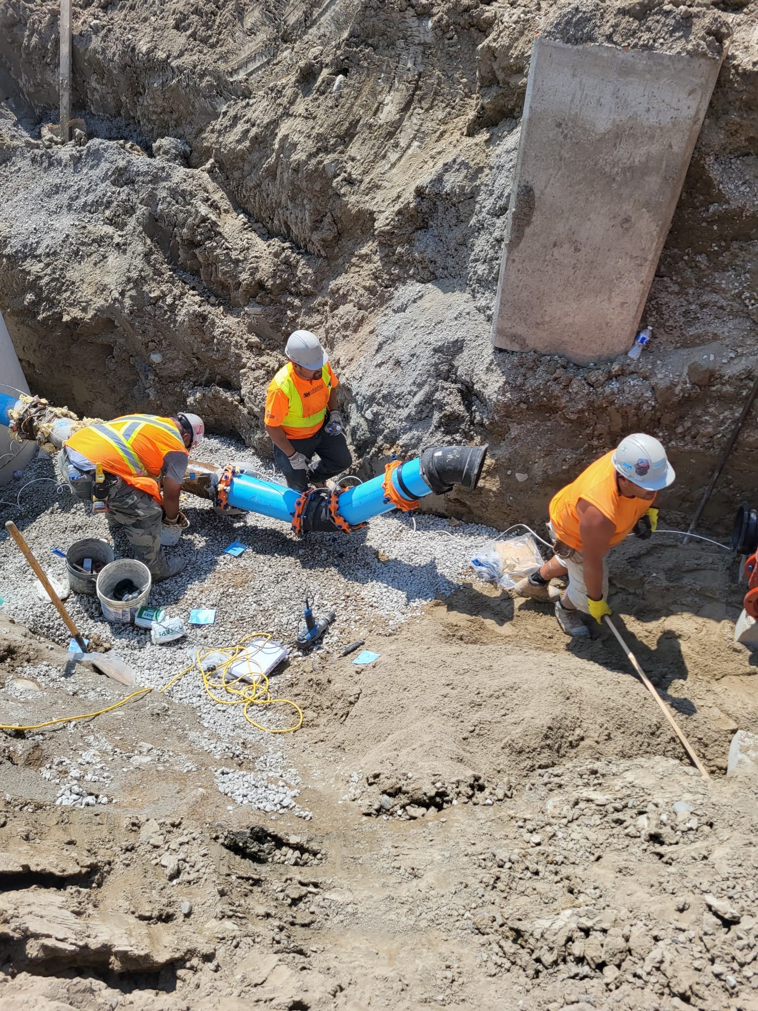 Three construction workers installing a large blue pipe in a trench at a construction site, wearing safety gear including helmets and reflective vests.
