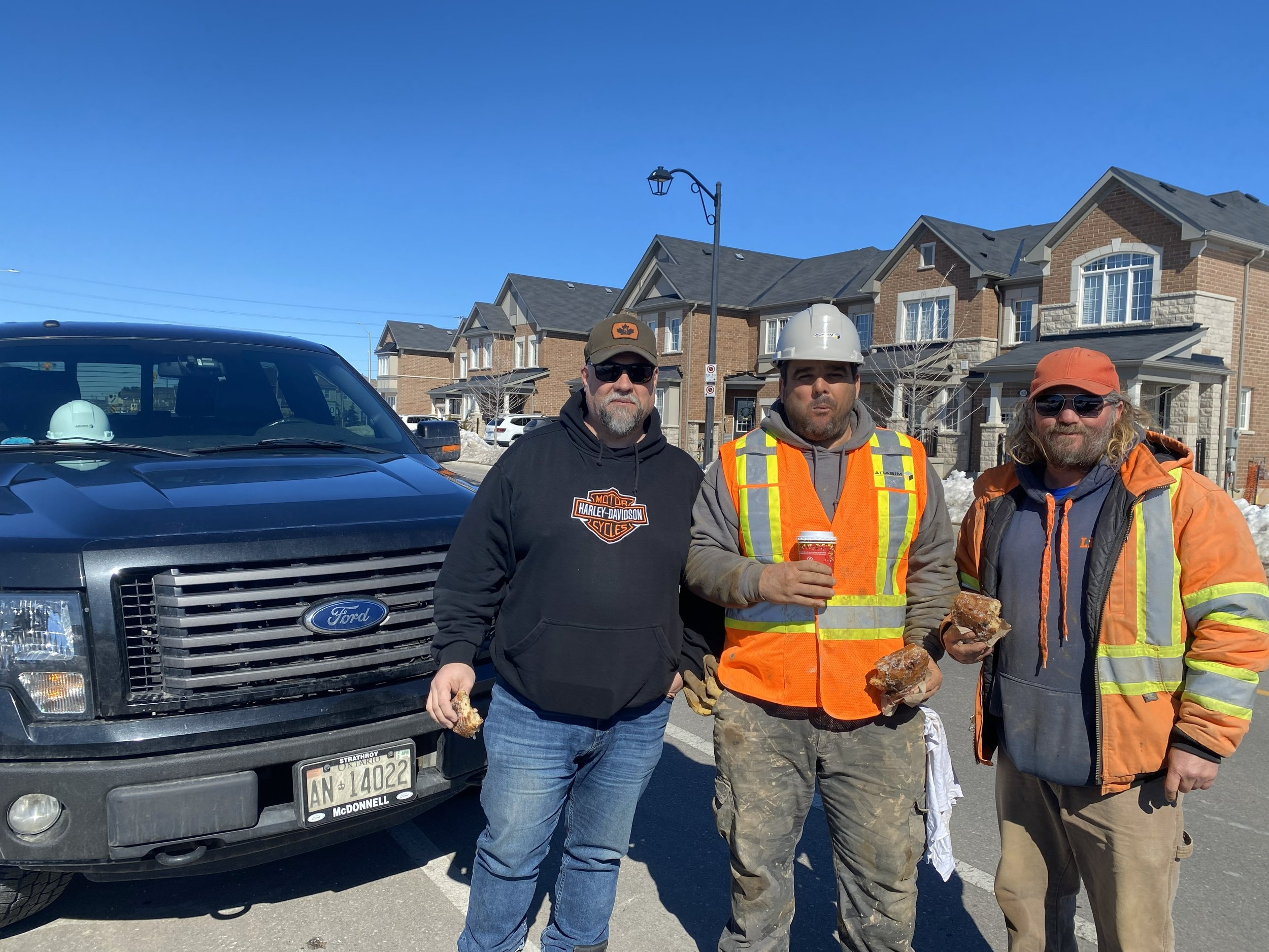 Three men standing in a residential neighborhood, three are holding donuts, one has a drink, all wearing work attire, with a back-up truck and houses in the background.