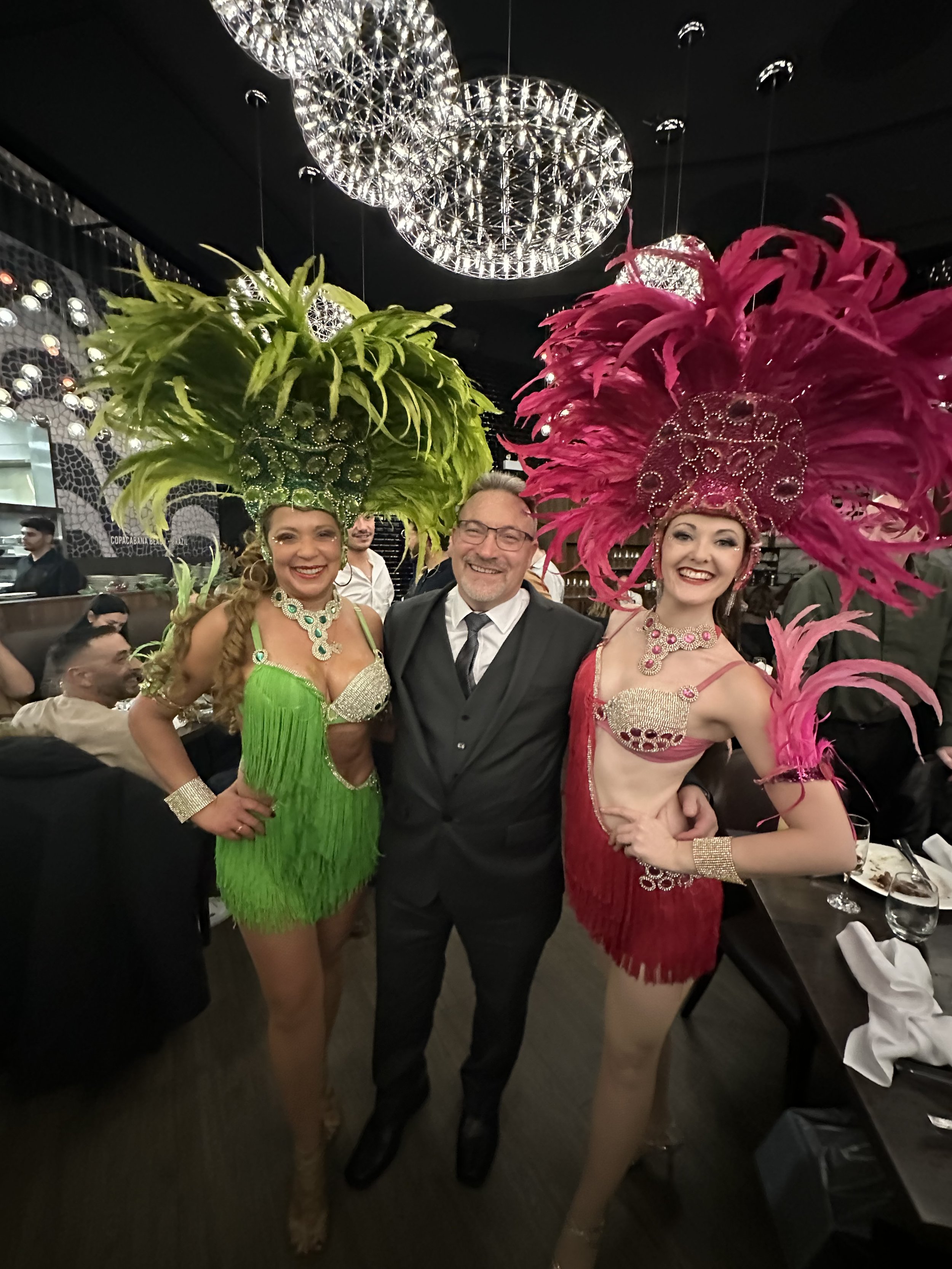 Two women in colorful feathers and sequined costumes with a man in a suit at a festive event or celebration.